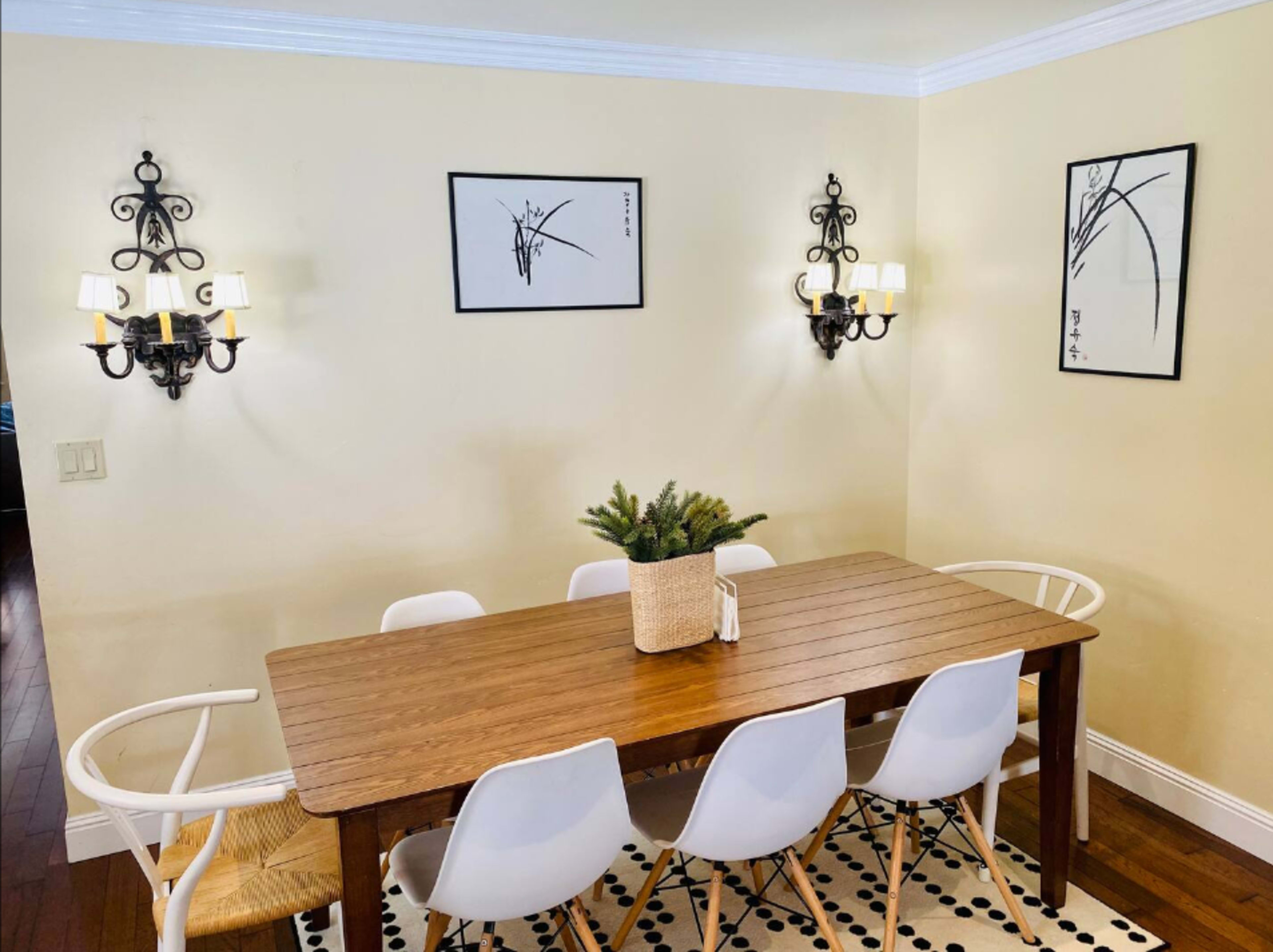 A rectangular wooden dining table is surrounded by white chairs in a well-lit dining area with two wall sconces and framed artwork on the walls.