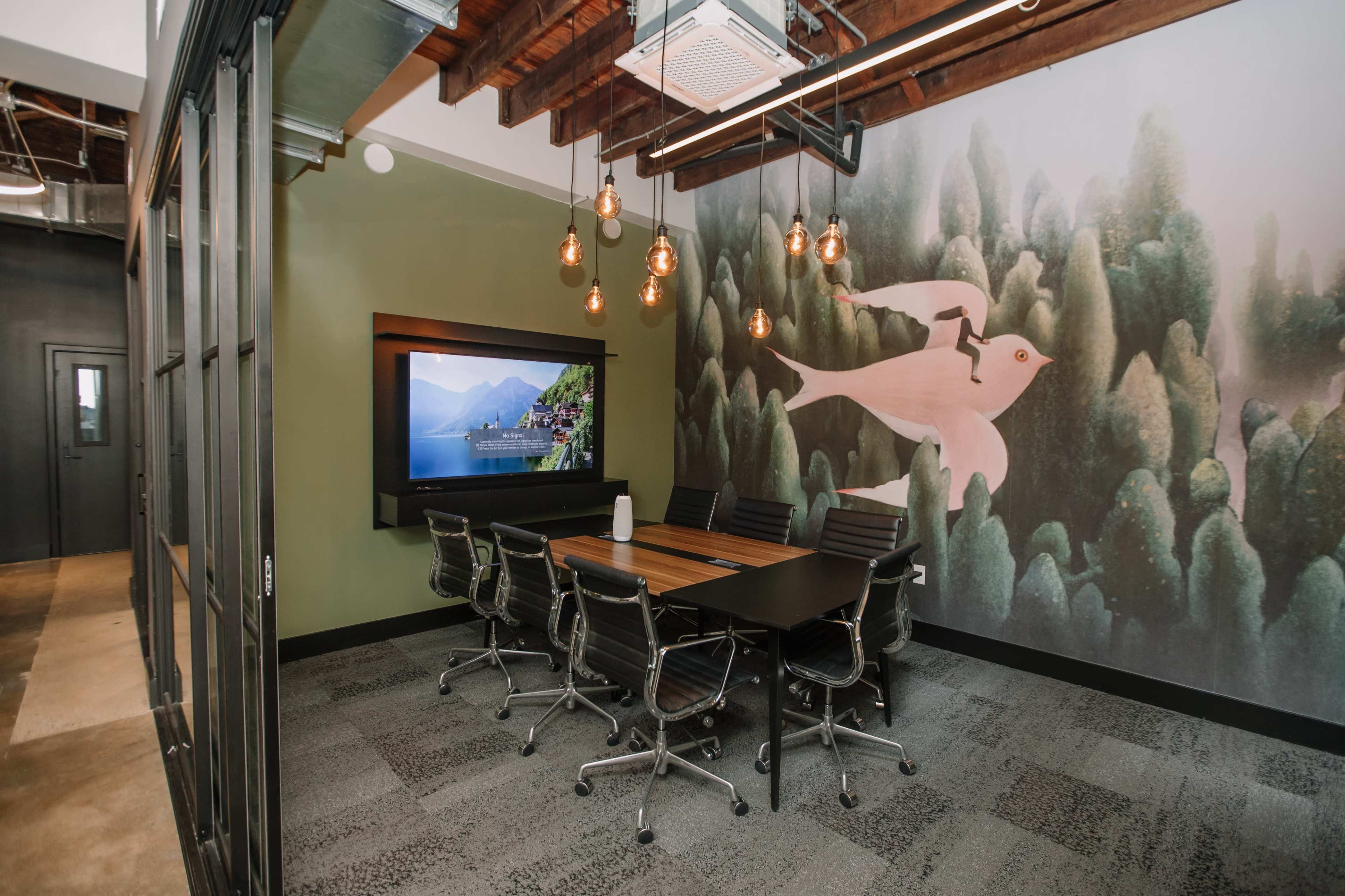 The image shows a modern conference room with a large wall-mounted screen, a wooden table surrounded by black office chairs, and a mural of mountains and a bird on one wall, illuminated by pendant lights.
