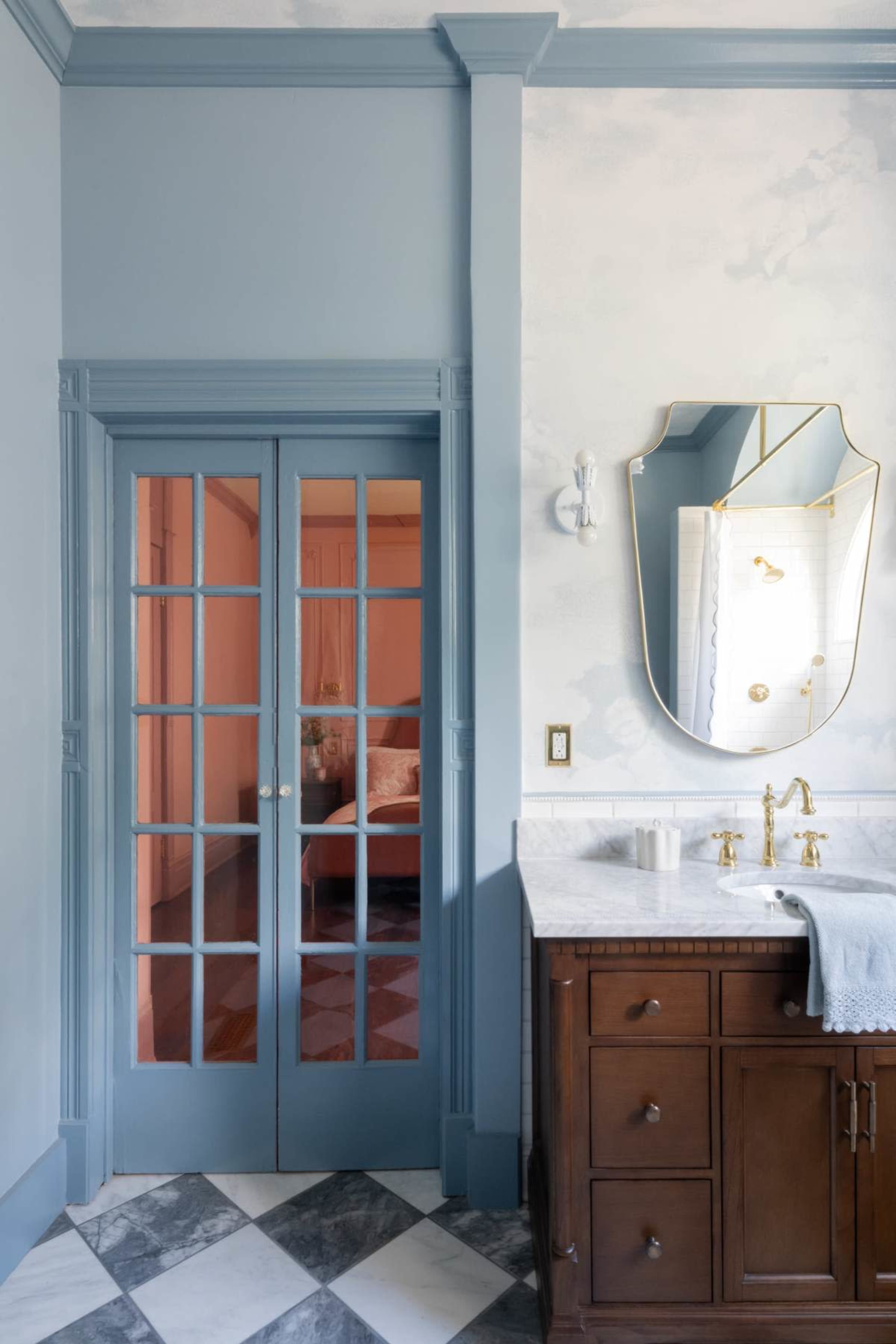 The image shows a bathroom with a blue and white color scheme, featuring a set of double glass doors, a large mirror, and a brown wooden vanity with marble countertops.