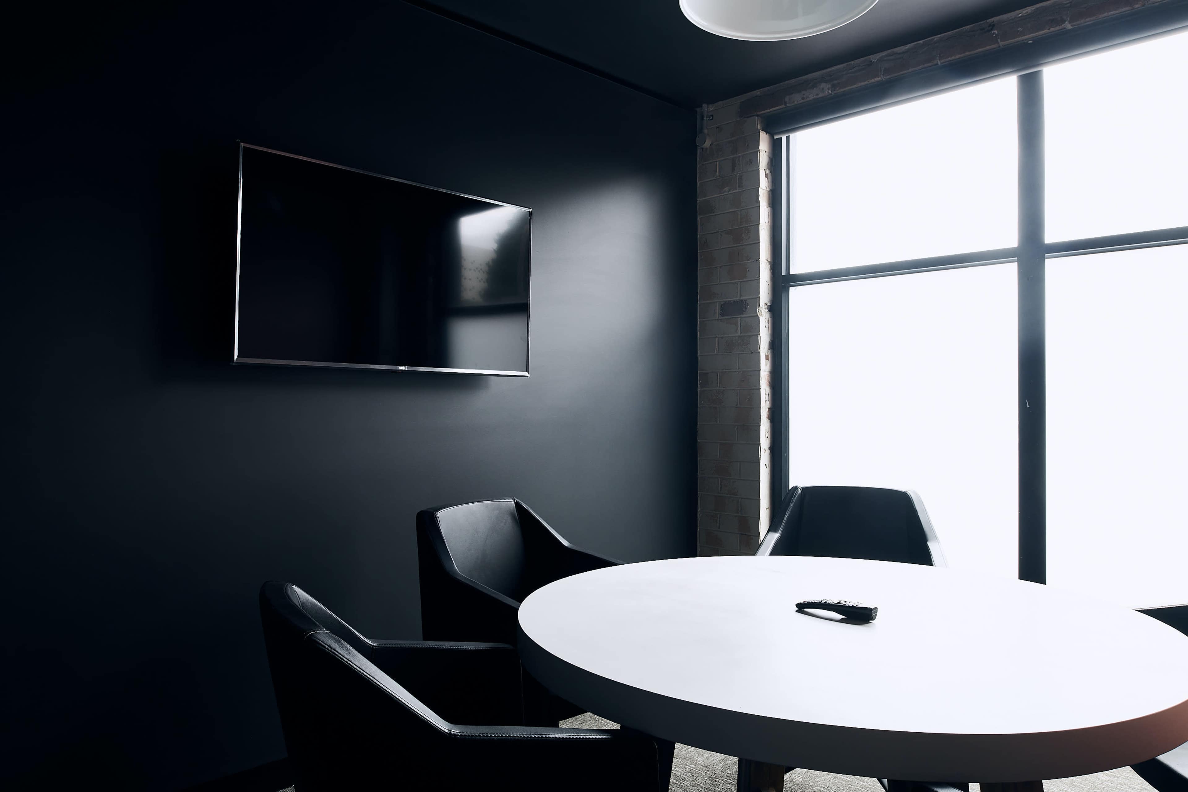The image shows a small, dark conference room with a round table, three chairs, and a wall-mounted television beside a large window.
