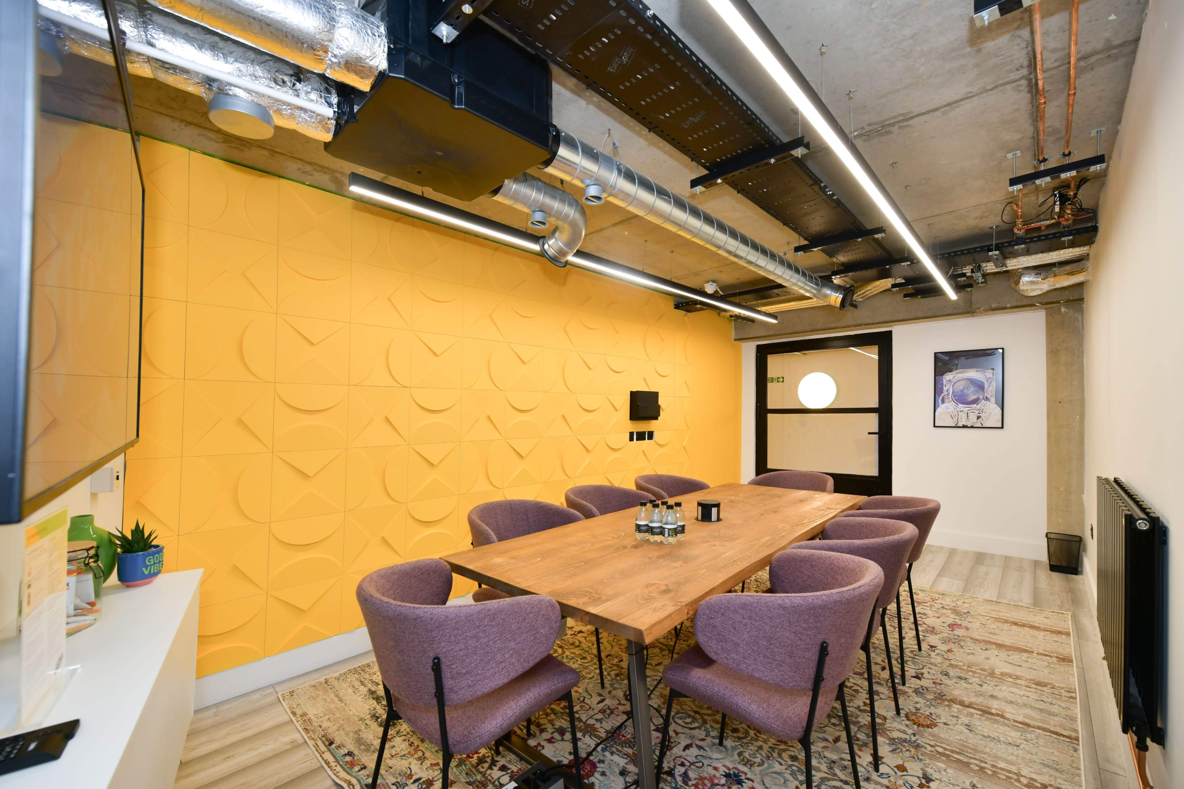 A modern meeting room with a long wooden table surrounded by purple upholstered chairs, complemented by a bright yellow textured wall and exposed ceiling elements.