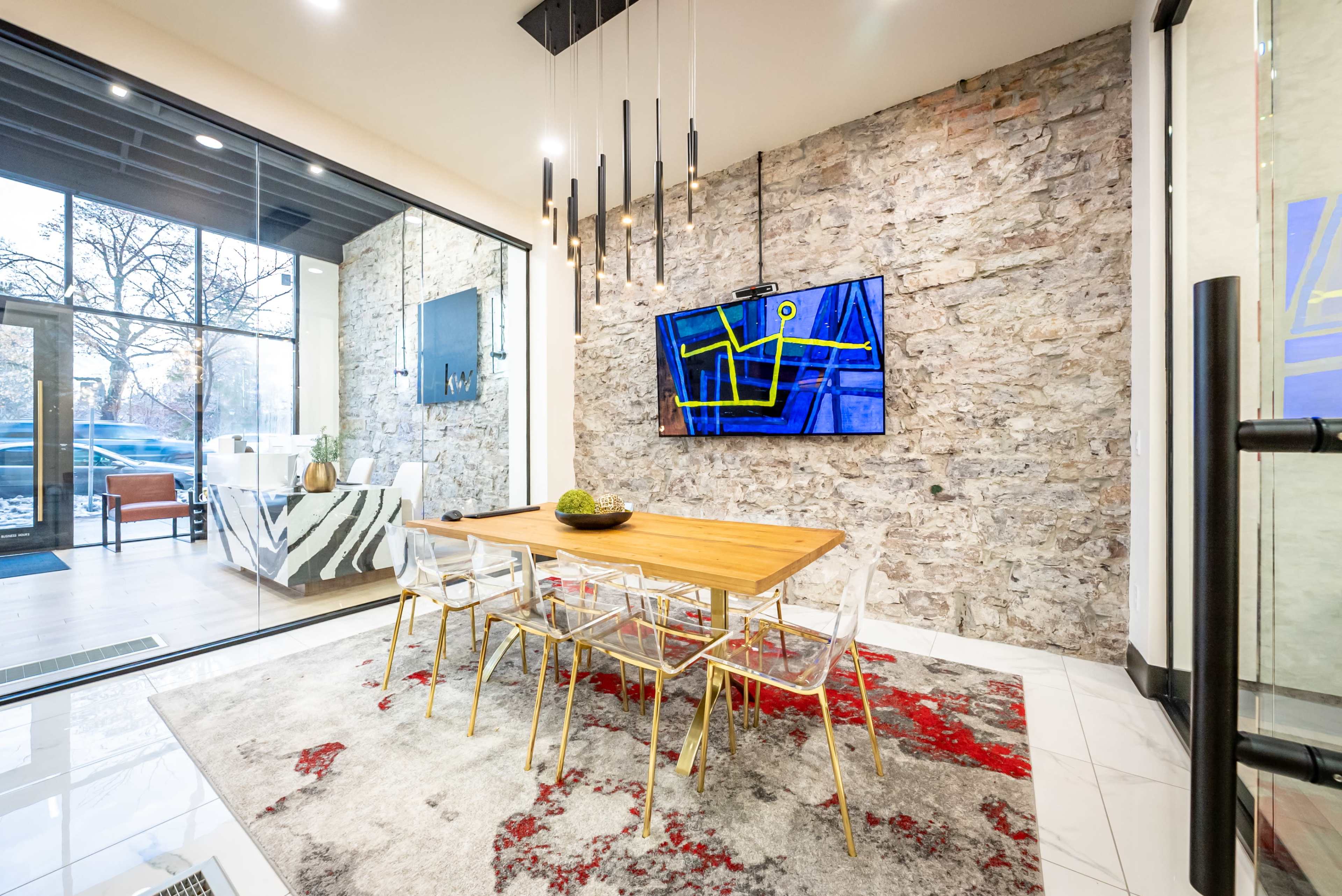 A modern dining area features a wooden table surrounded by clear chairs, with a large television displaying art on a stone wall backdrop.