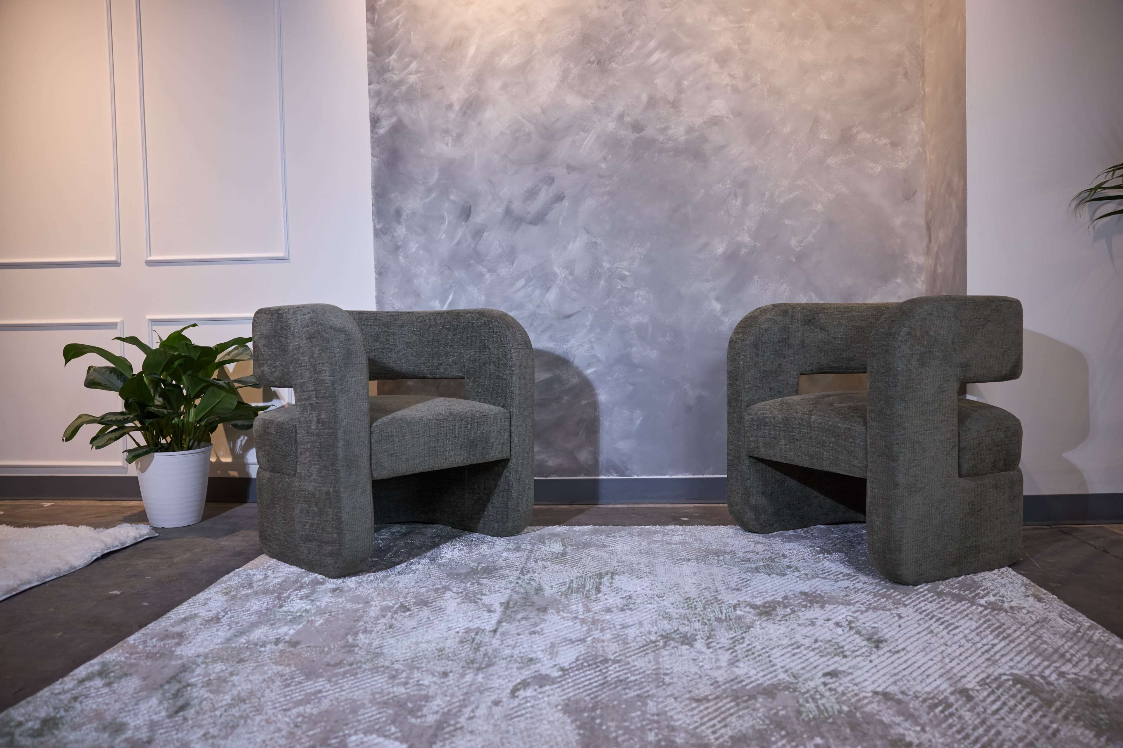 Two modern, upholstered chairs with a curved design are positioned facing each other on a patterned rug in a minimalist interior space.