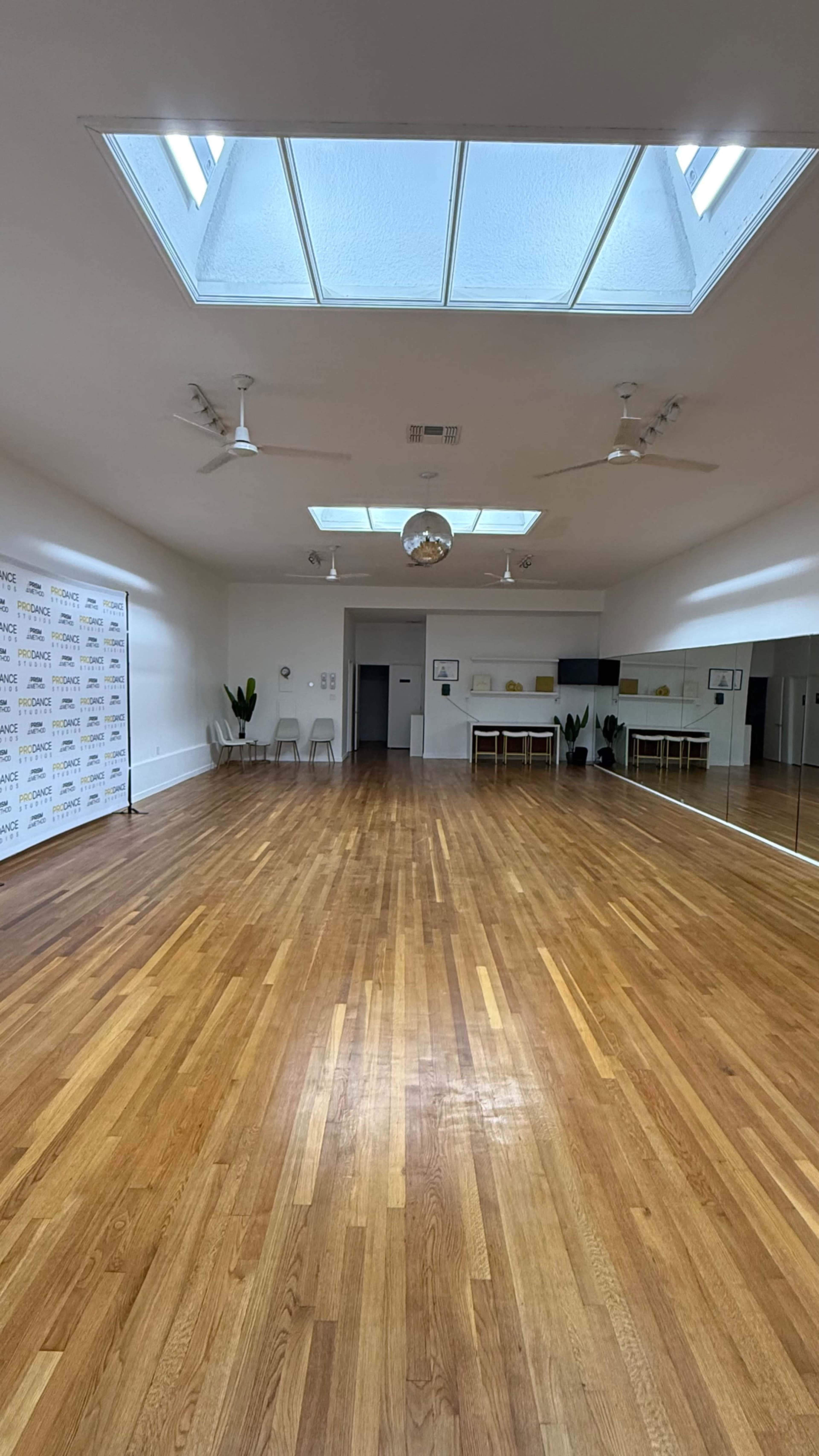 The image shows an empty wooden-floored studio with large windows and a mirror on one wall.