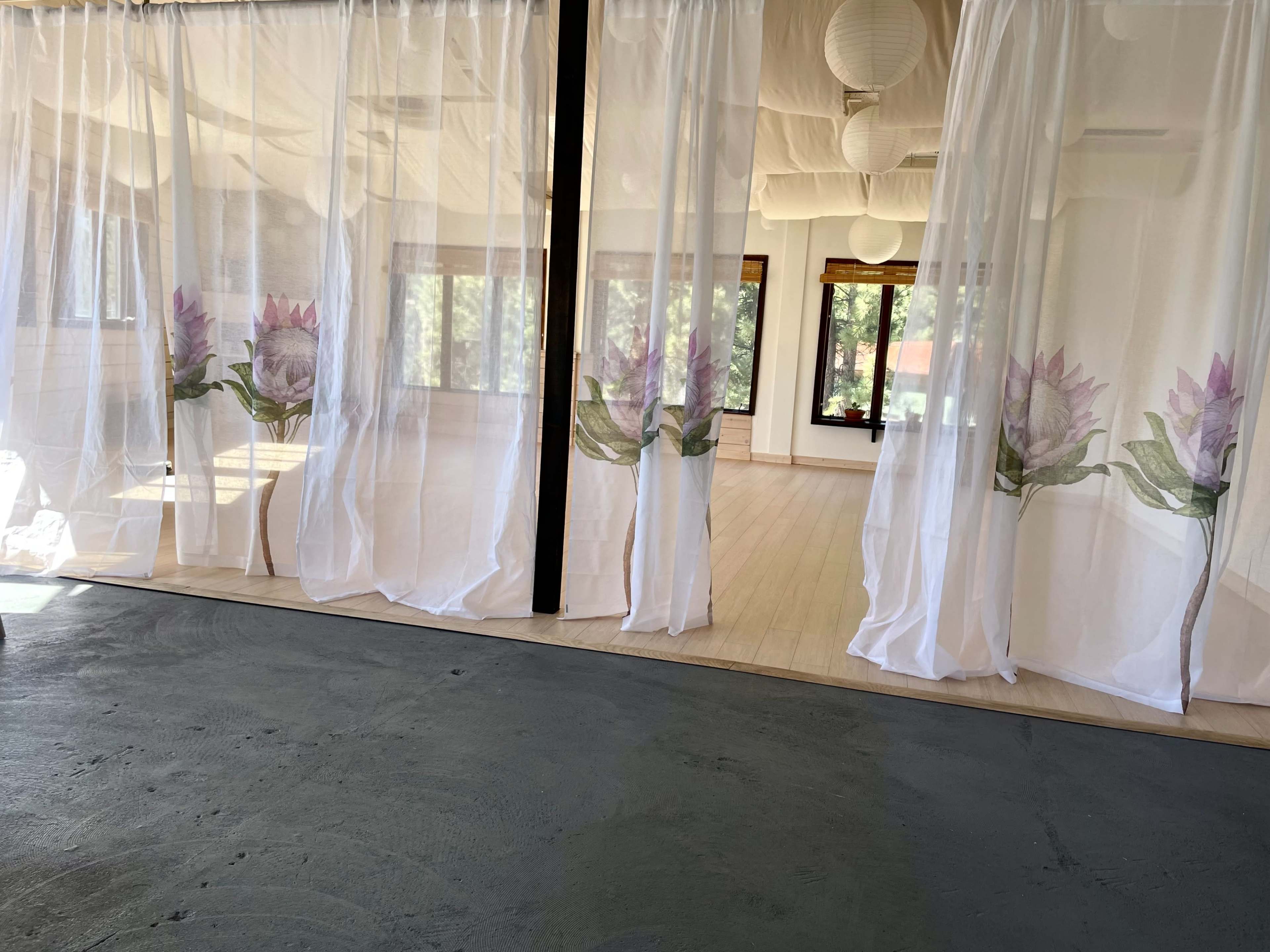 A spacious room divided by sheer curtains featuring floral designs, with large windows allowing natural light to enter.