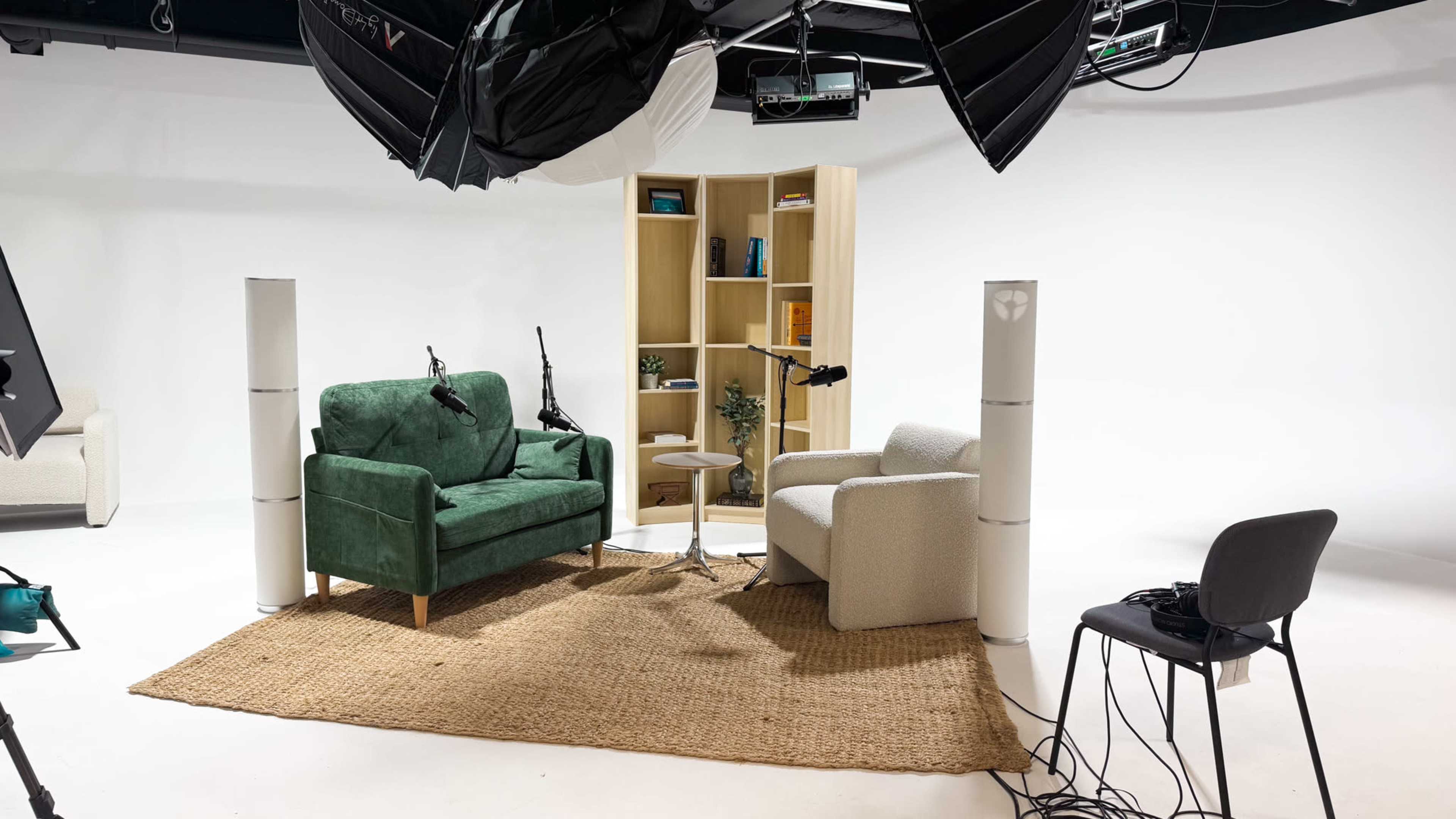 The image shows a minimalist studio setup featuring two chairs, a small table, a bookshelf with decor items, and professional lighting equipment.