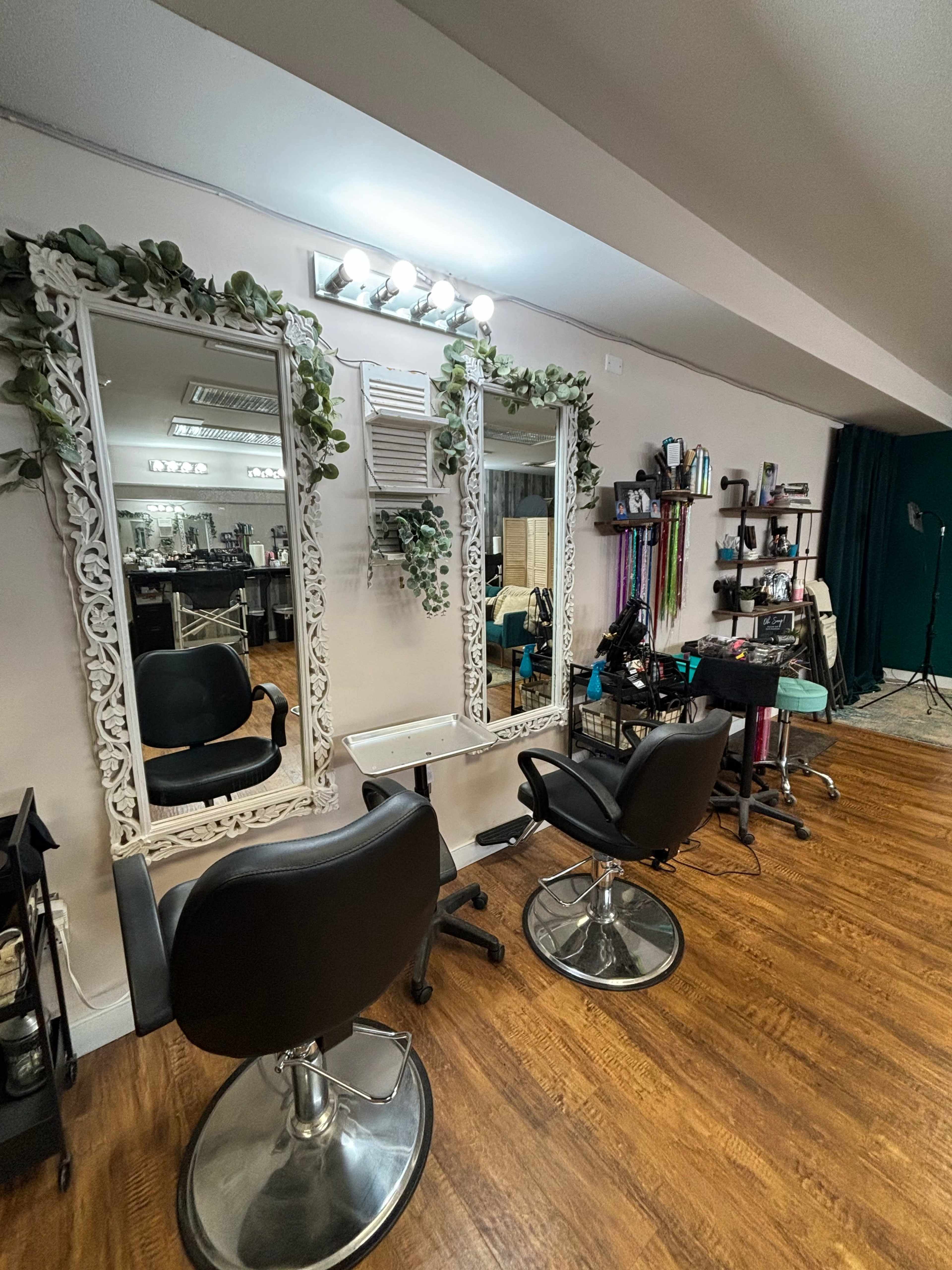 A hair salon with two mirrored stations, each equipped with a chair, surrounded by decorative plants and various styling tools on shelves.