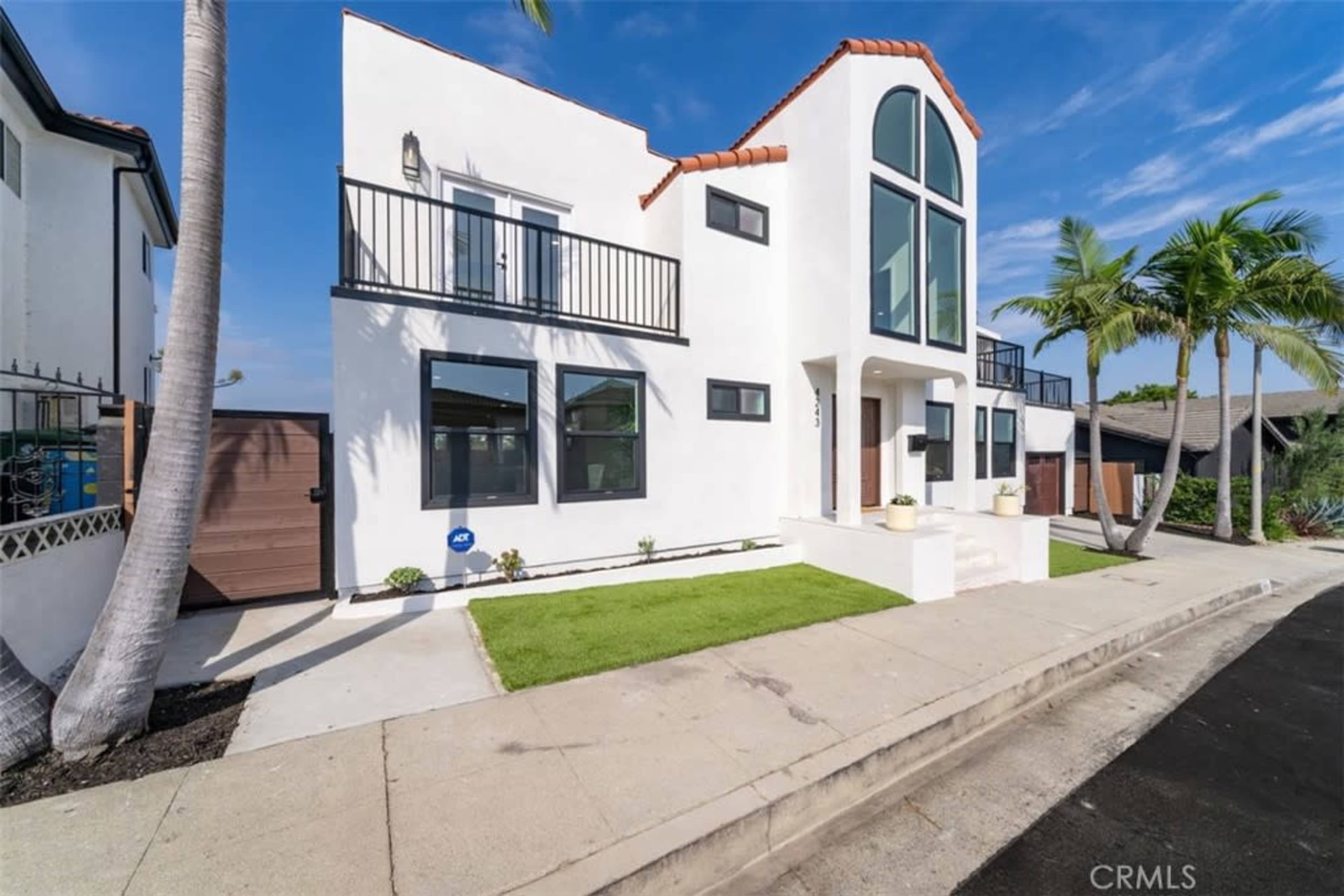 The image shows a modern two-story house with a distinctive peaked roof, large windows, and palm trees in front, located on a residential street.
