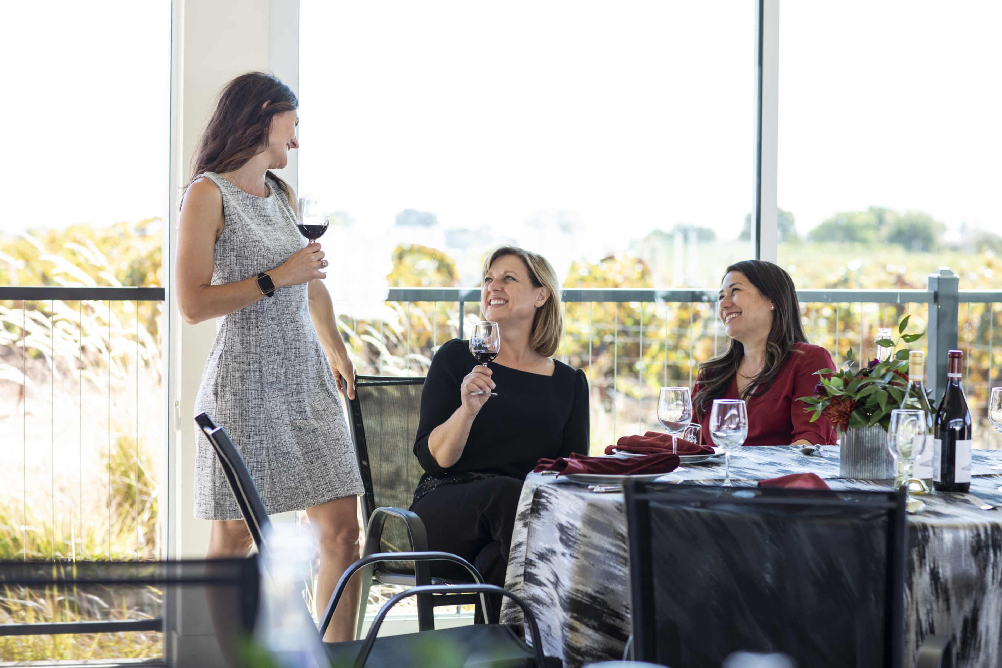 Three women are seated at a dining table in a well-lit room, while another woman stands nearby holding a glass of wine.