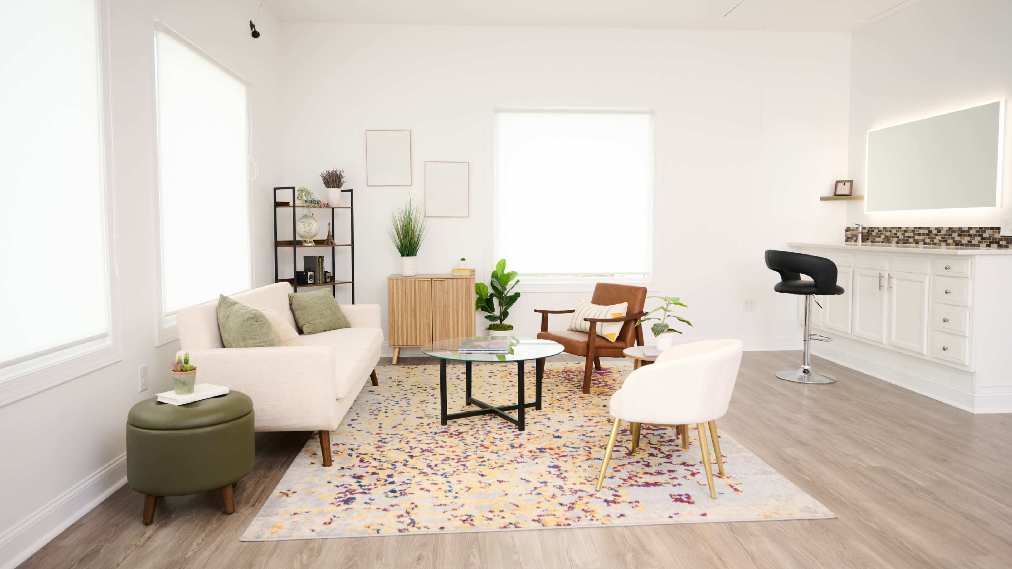 The image shows a bright, modern living room featuring a light sofa, a round coffee table, a small dining area, and a rug with purple and yellow patterns.