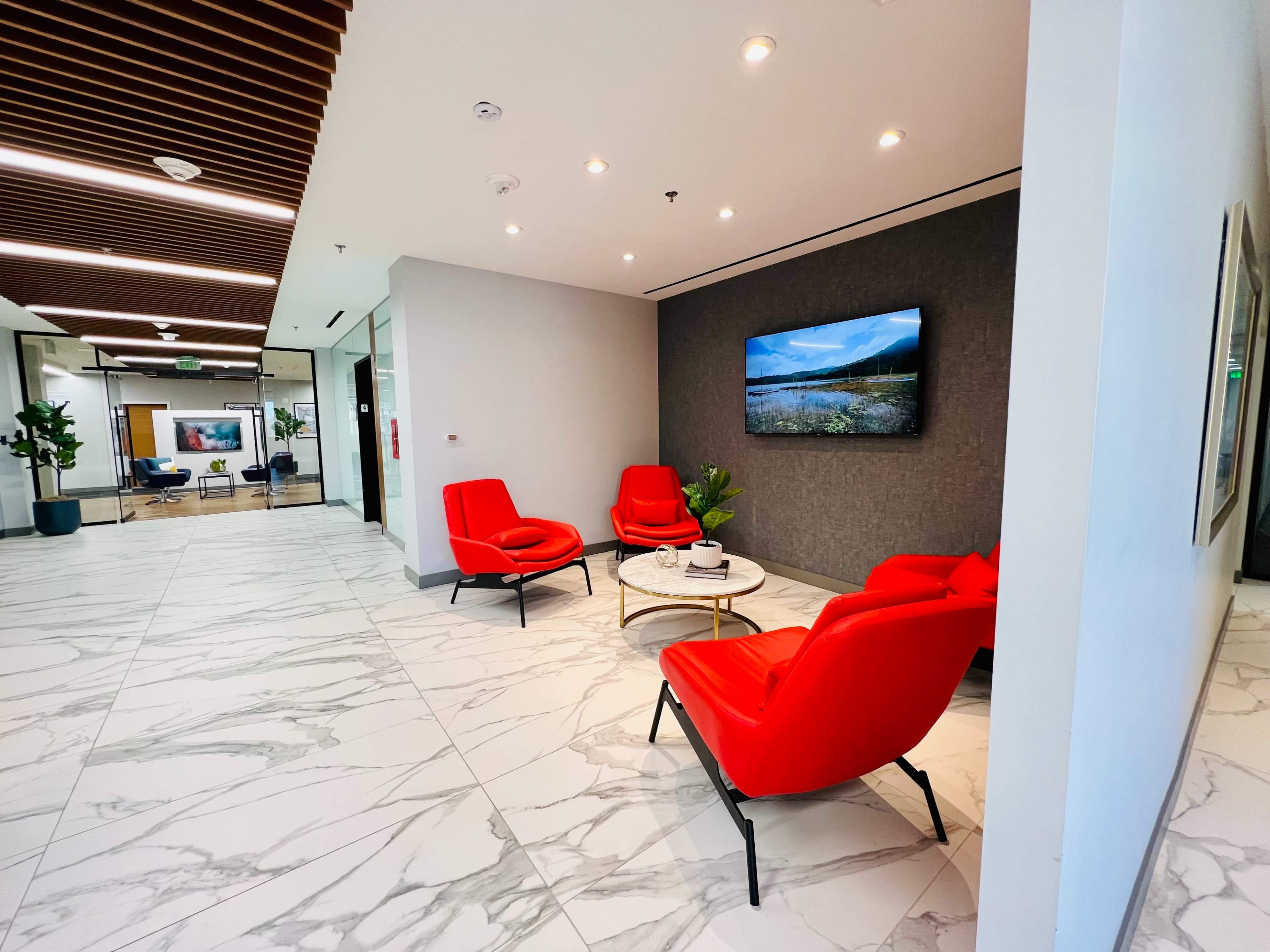 A modern lobby area with red chairs surrounding a white coffee table, a textured wall displaying a landscape on a flat-screen TV, and marble flooring.