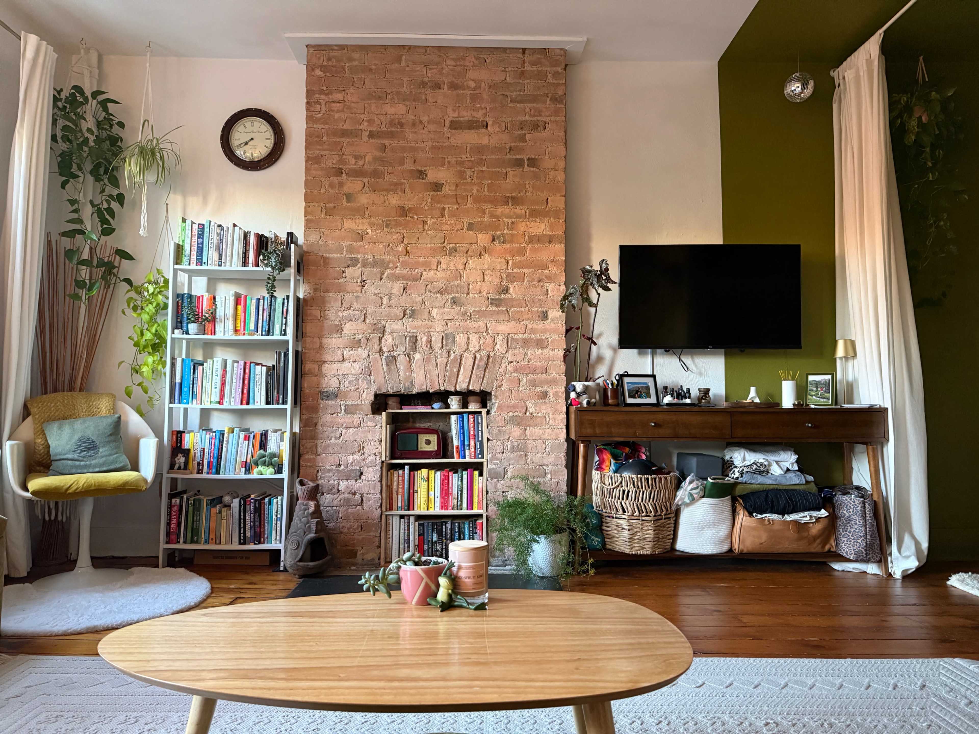 The room features a brick fireplace, a bookshelf filled with books, a TV mounted on the wall, and a coffee table in front, surrounded by plants and decorative items.
