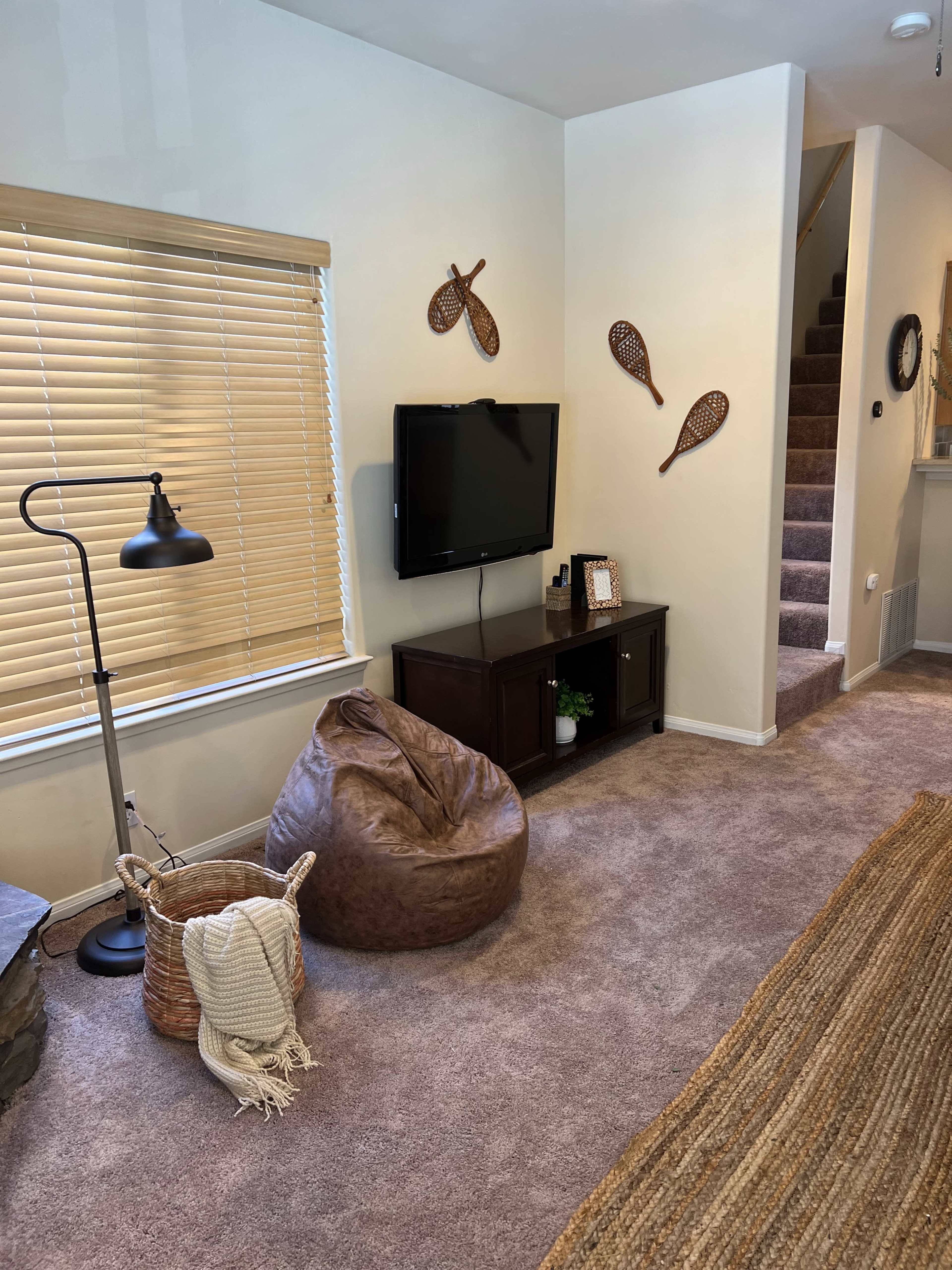 The room features a television mounted on a wall opposite a beige bean bag chair, with a decorative lamp and a woven basket nearby, set in a space with light-colored walls and carpet.