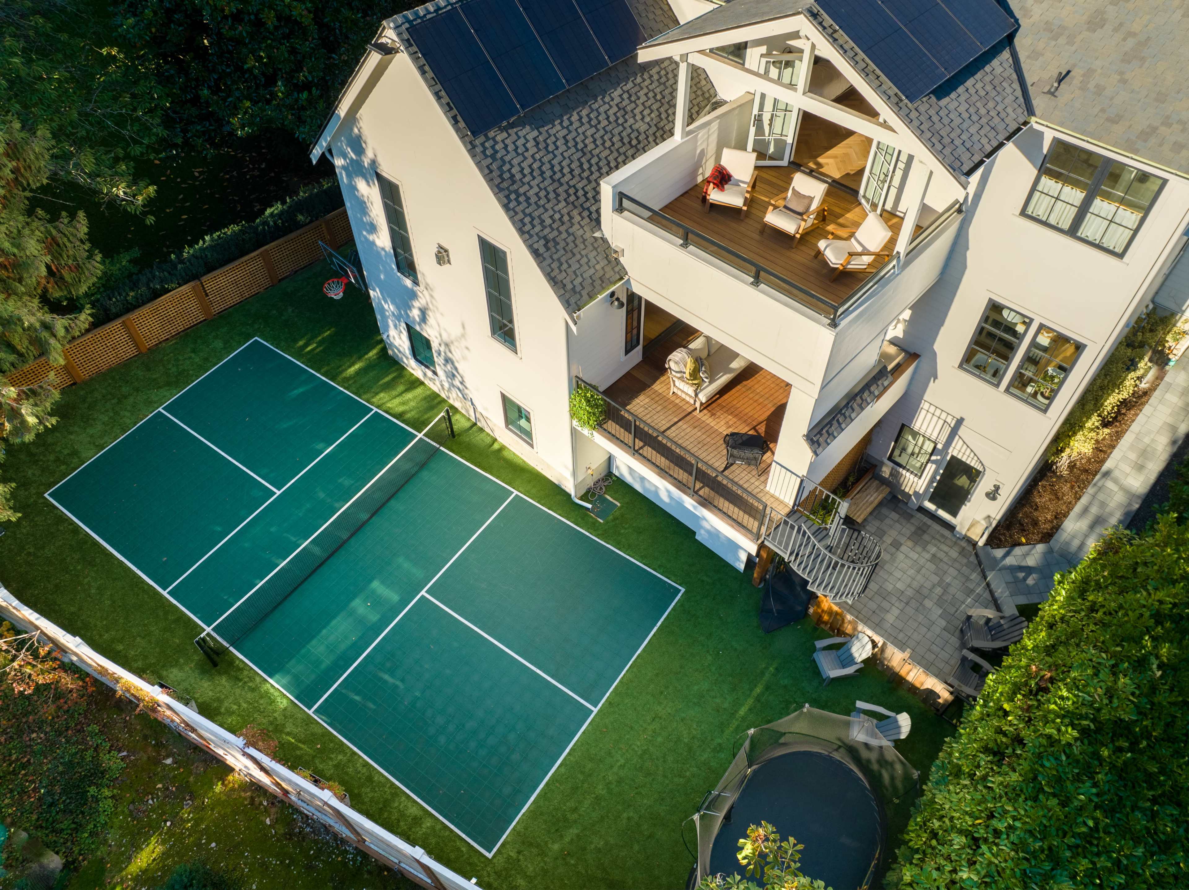 The image shows a two-story house with solar panels on the roof, a balcony with seating, and a green tennis court in the backyard.