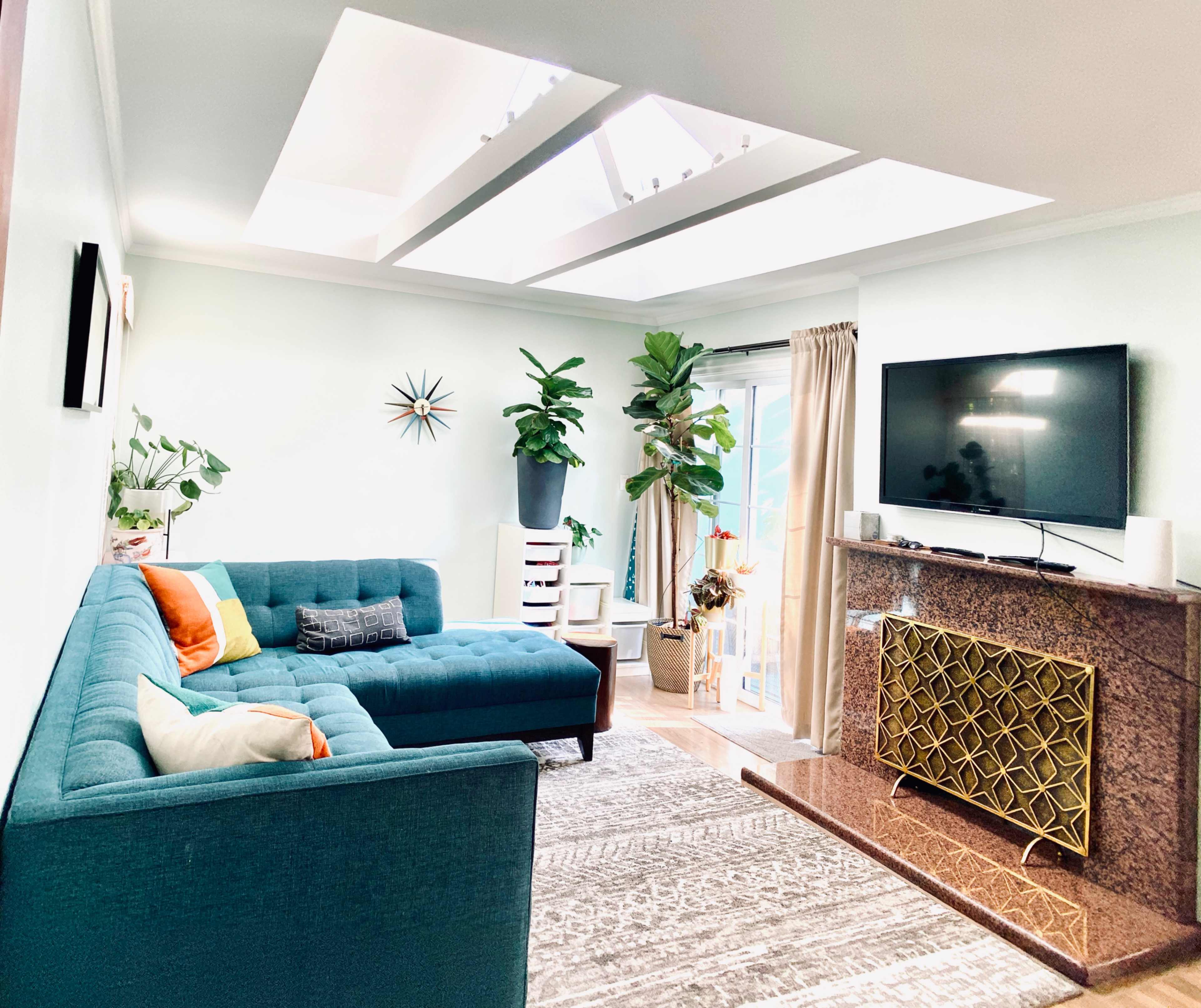 A bright living room featuring a teal couch, a large television on a granite fireplace, and several plants near a skylight.