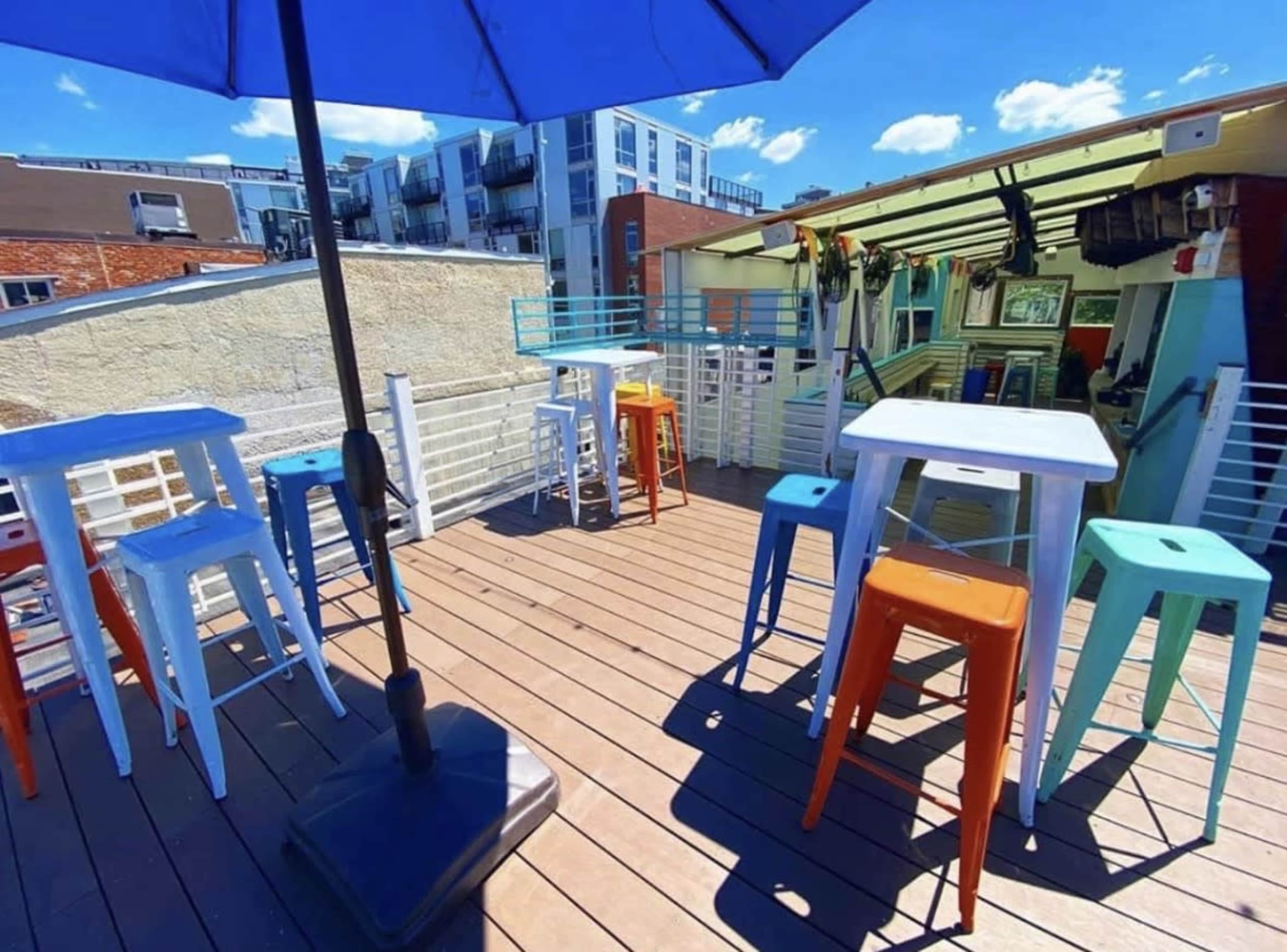 The image shows a rooftop bar area with colorful metal stools and tables under a blue umbrella.