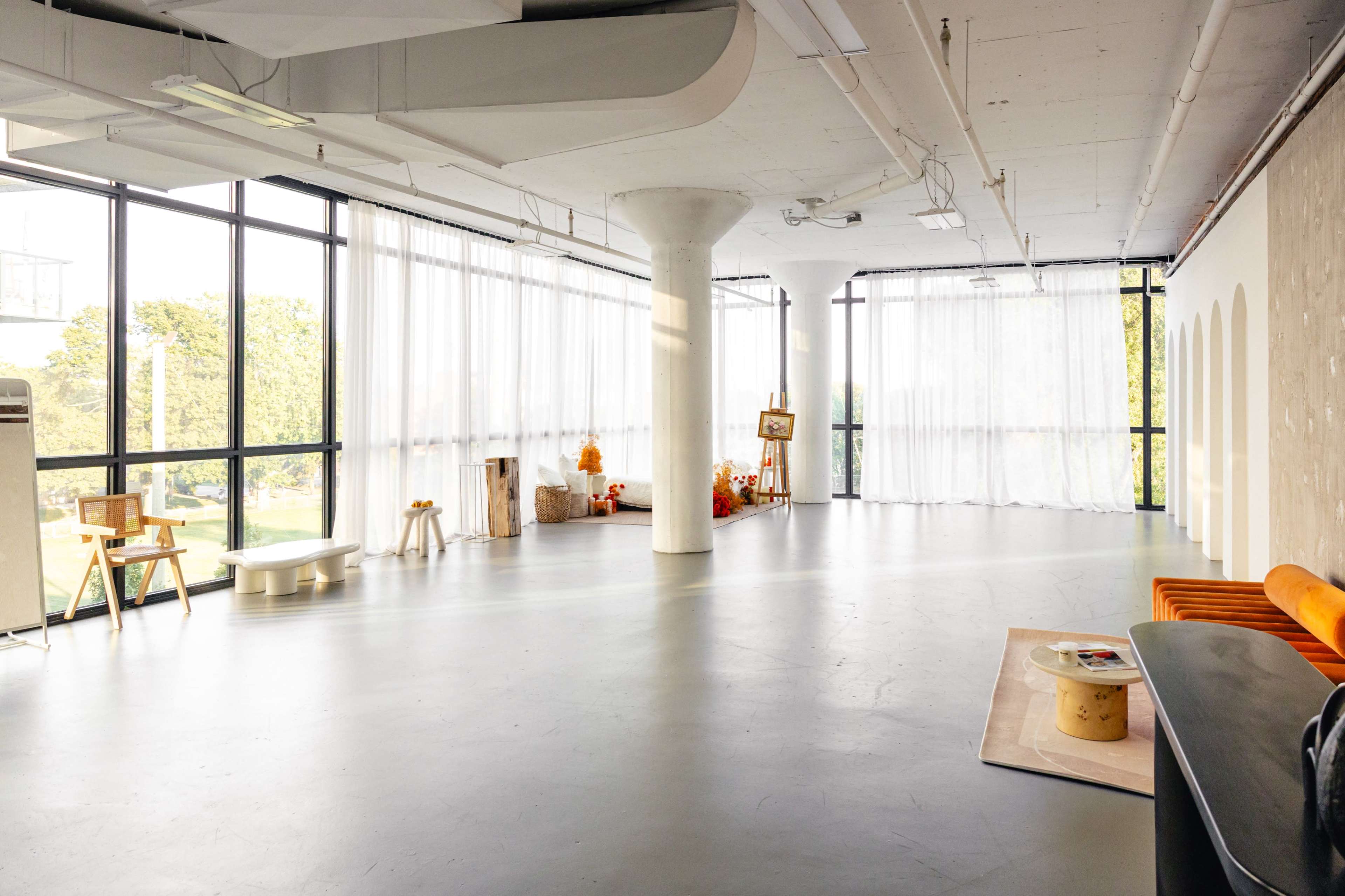 The image shows a spacious, well-lit interior room with large windows, minimalist furniture, and light-colored walls.