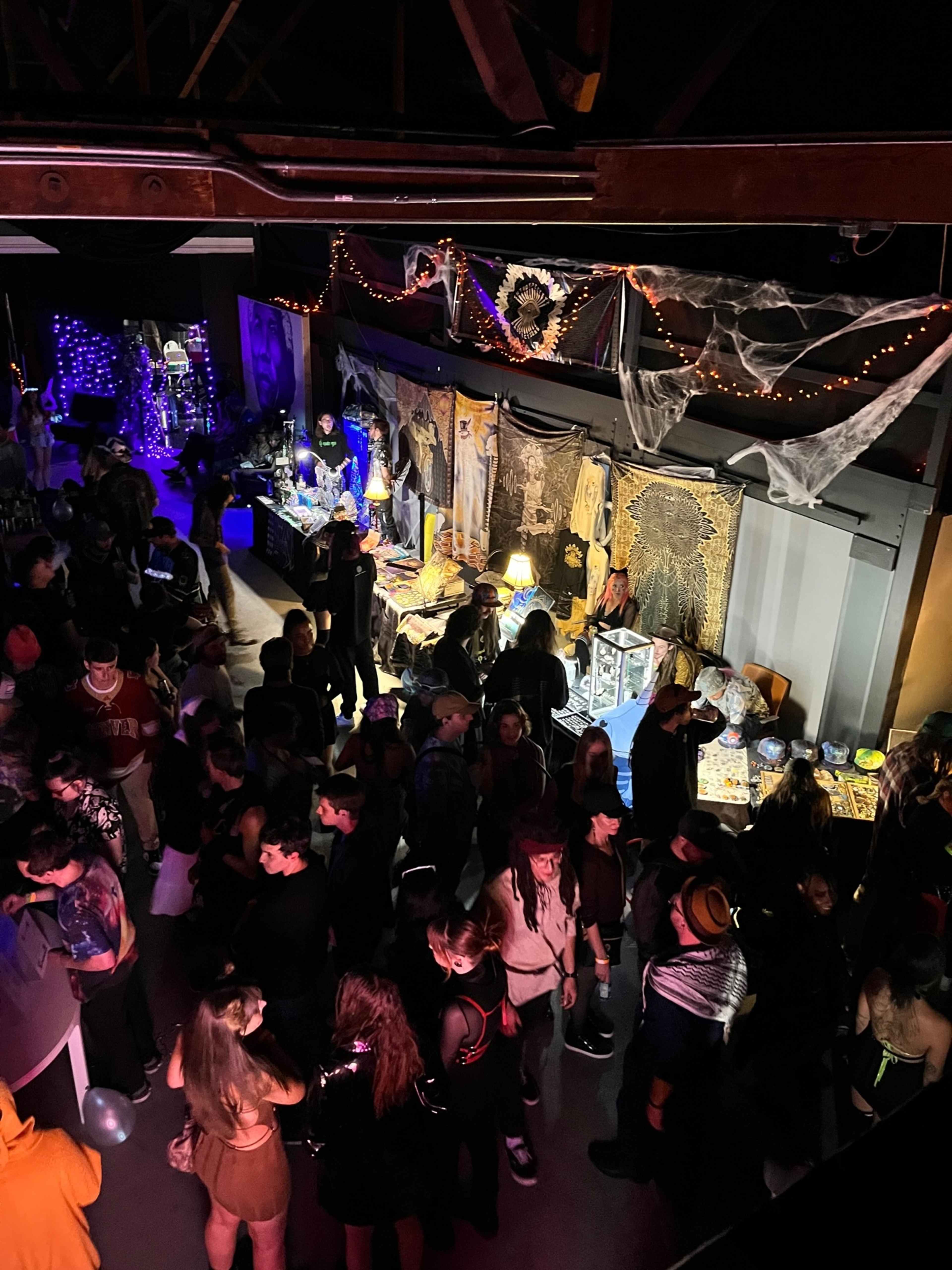 A crowd of people gathers in a dimly lit party venue decorated with spider webs and various props.