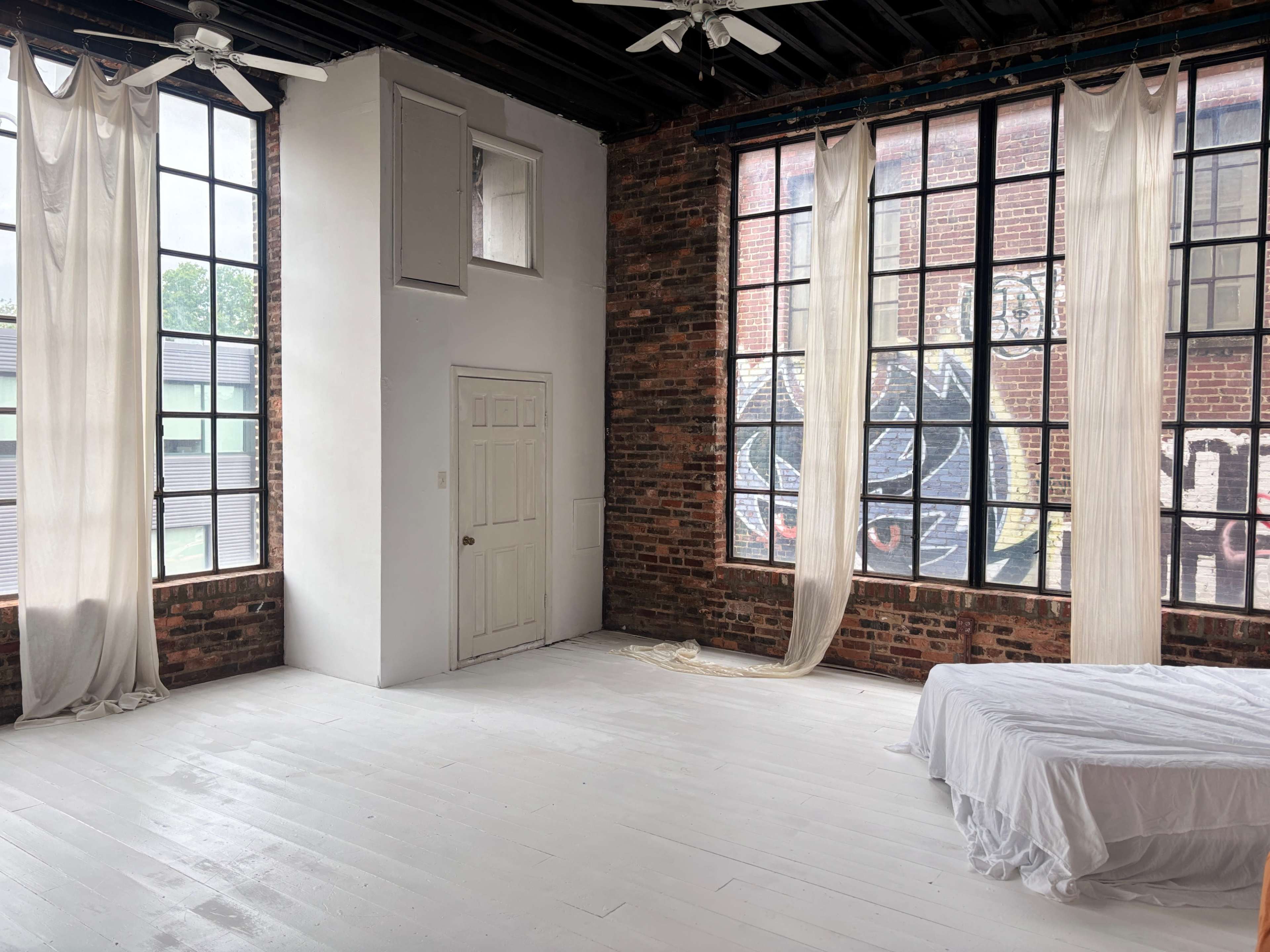 The interior features exposed brick walls, large windows with sheer curtains, and a minimalist bed in a bright, airy space.
