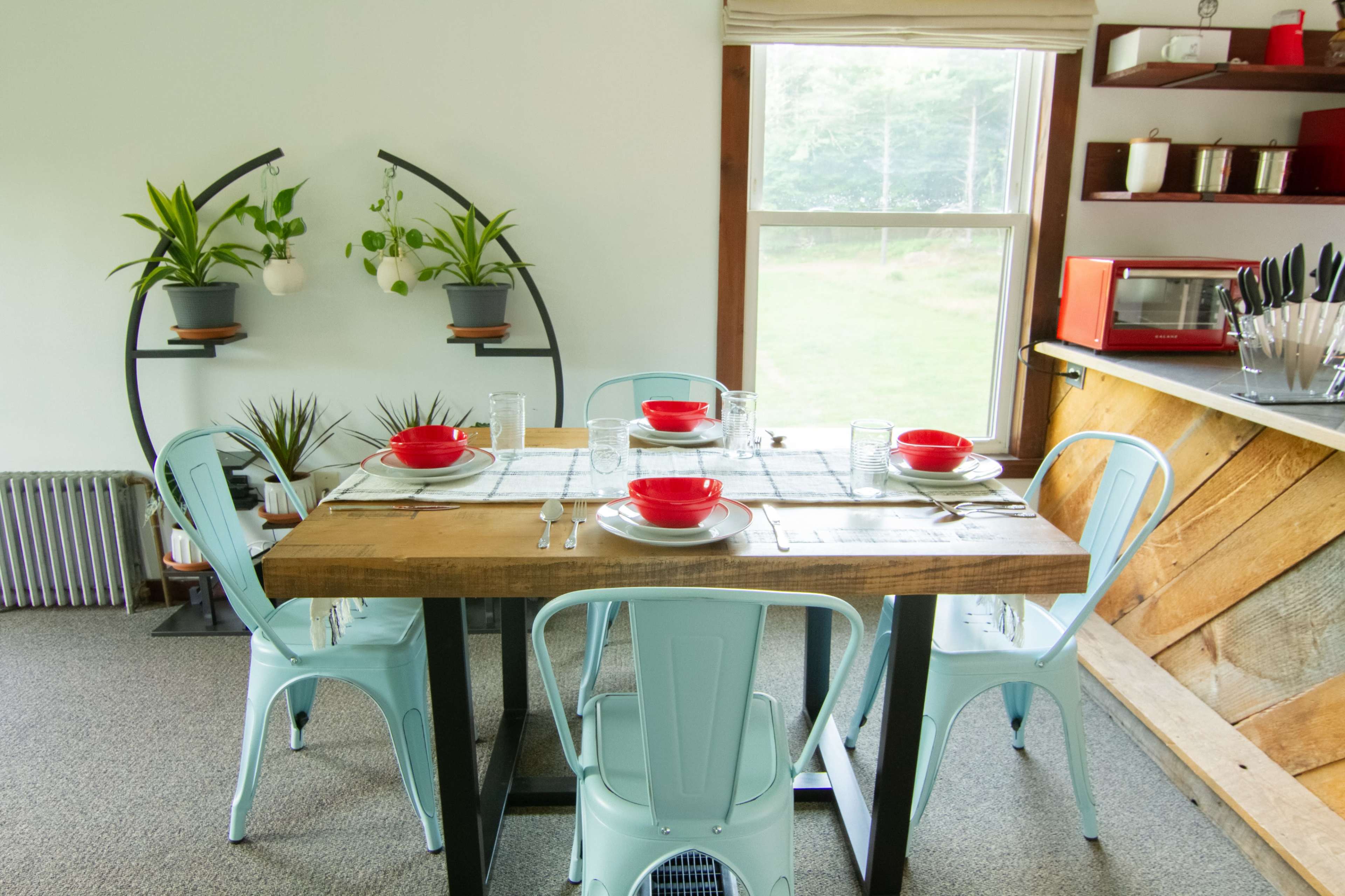 The image shows a dining area with a wooden table set for four, featuring blue metal chairs, red bowls, and green potted plants in a well-lit space.