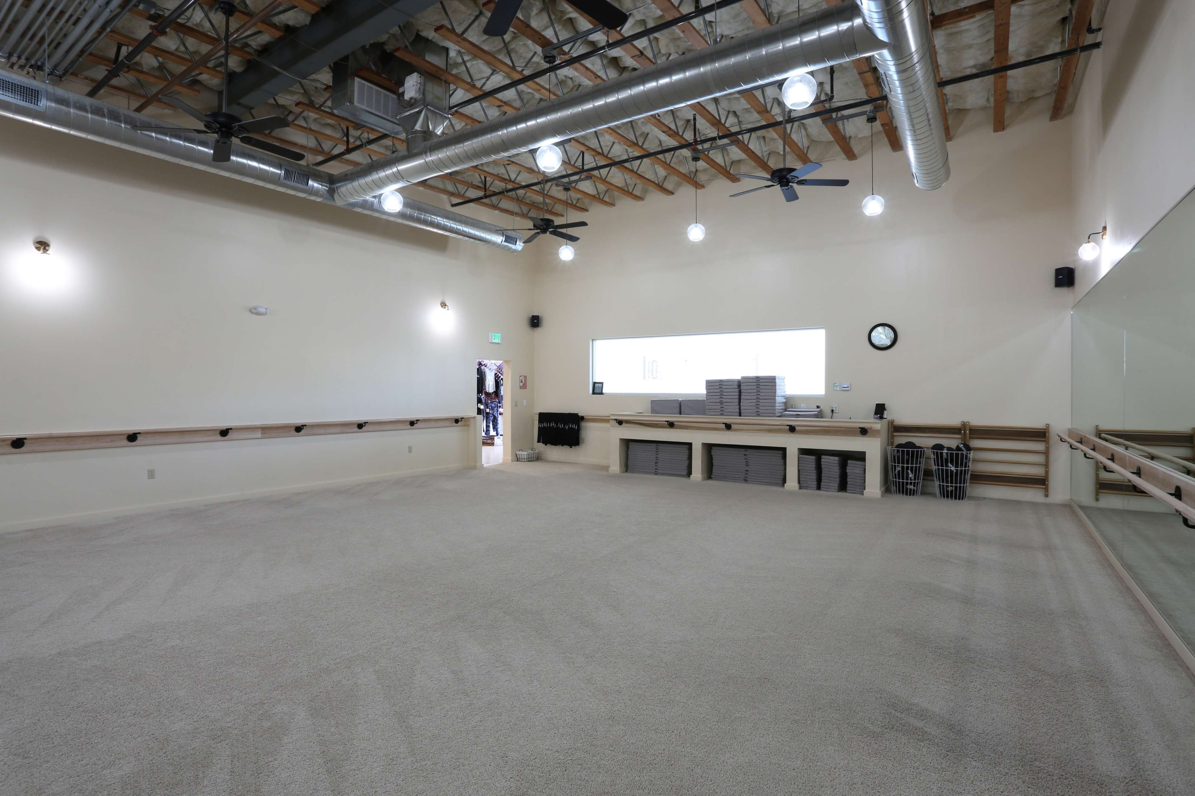 An empty studio space with carpeted flooring, wall-mounted ballet bars, and large mirrors, illuminated by overhead lights.