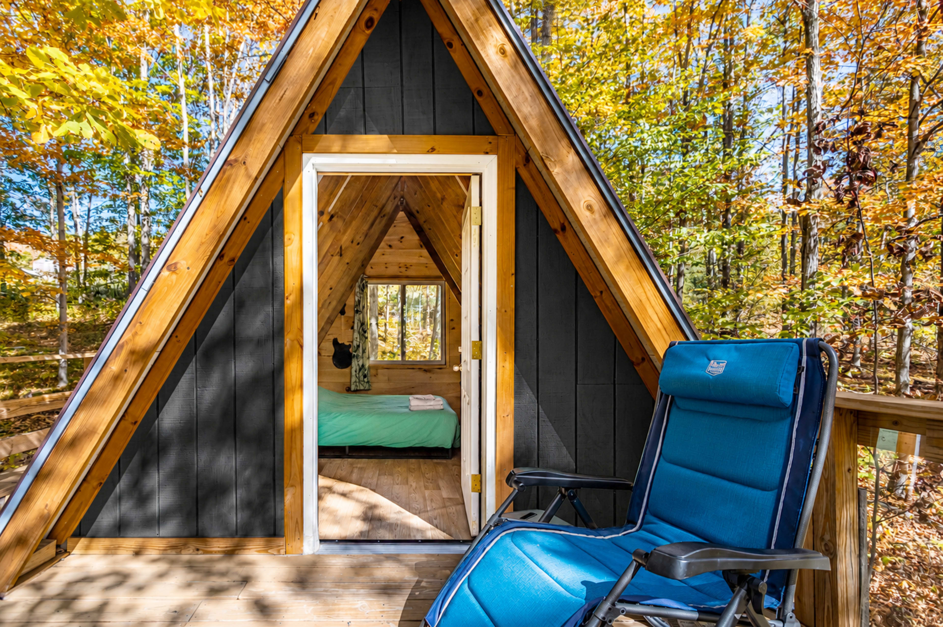 A wooden A-frame cabin with a door open to a bedroom and a blue chair on the porch surrounded by autumn foliage.