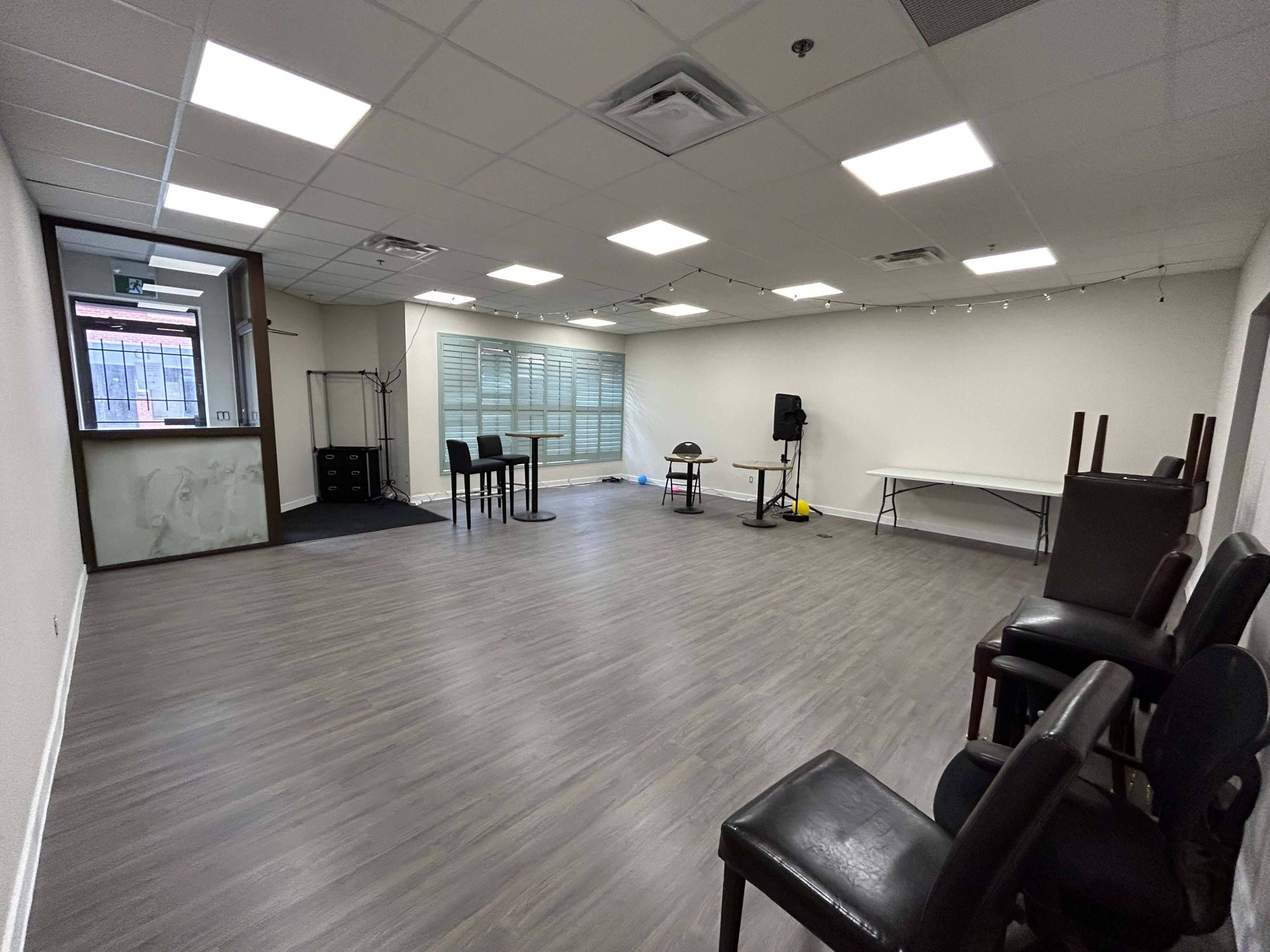 The image shows a spacious, empty room with wooden flooring, a few tables and chairs, and large windows allowing natural light to enter.