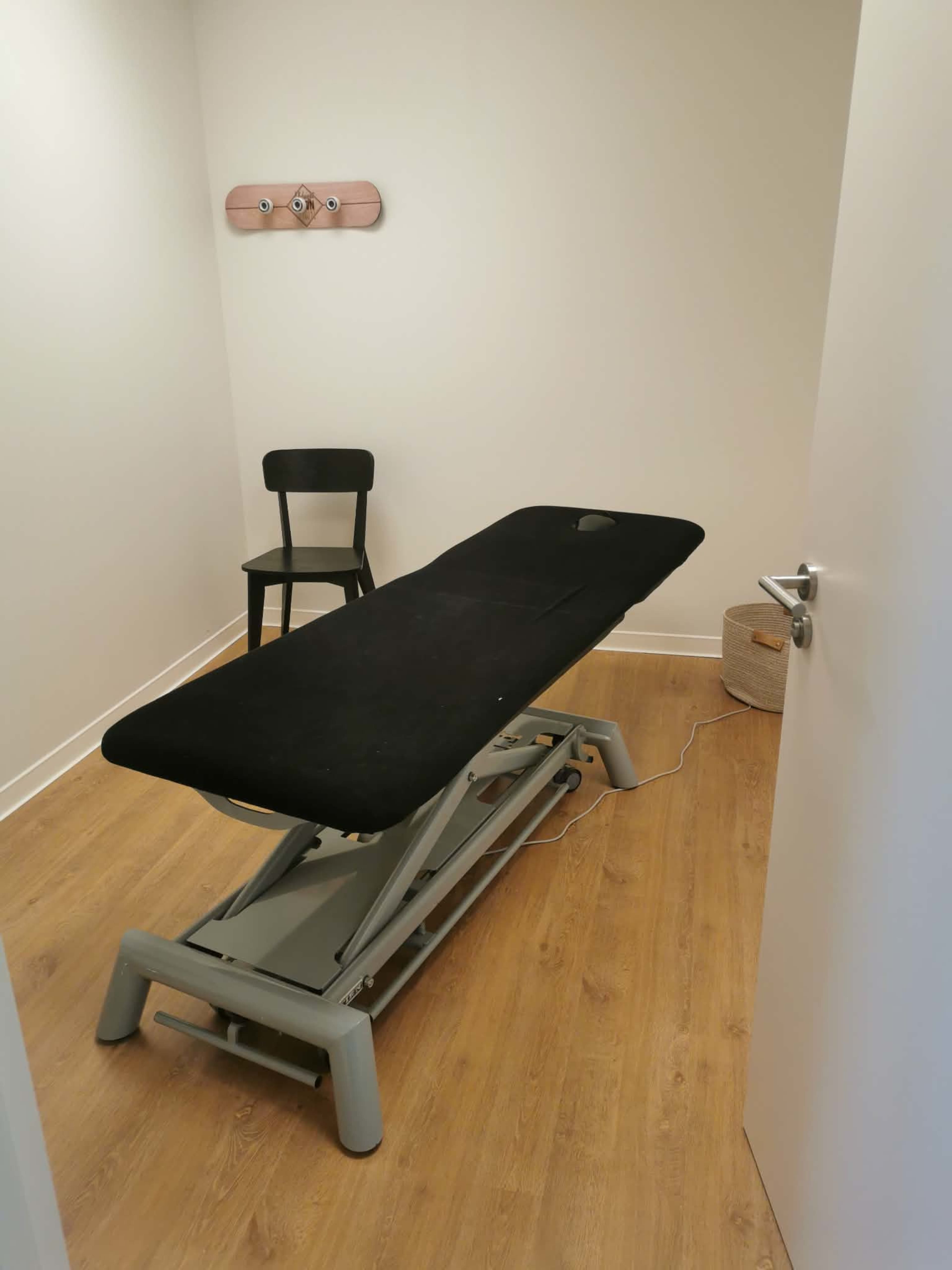 The image shows a small treatment room with a black massage table, a chair, and a light-colored wooden floor.