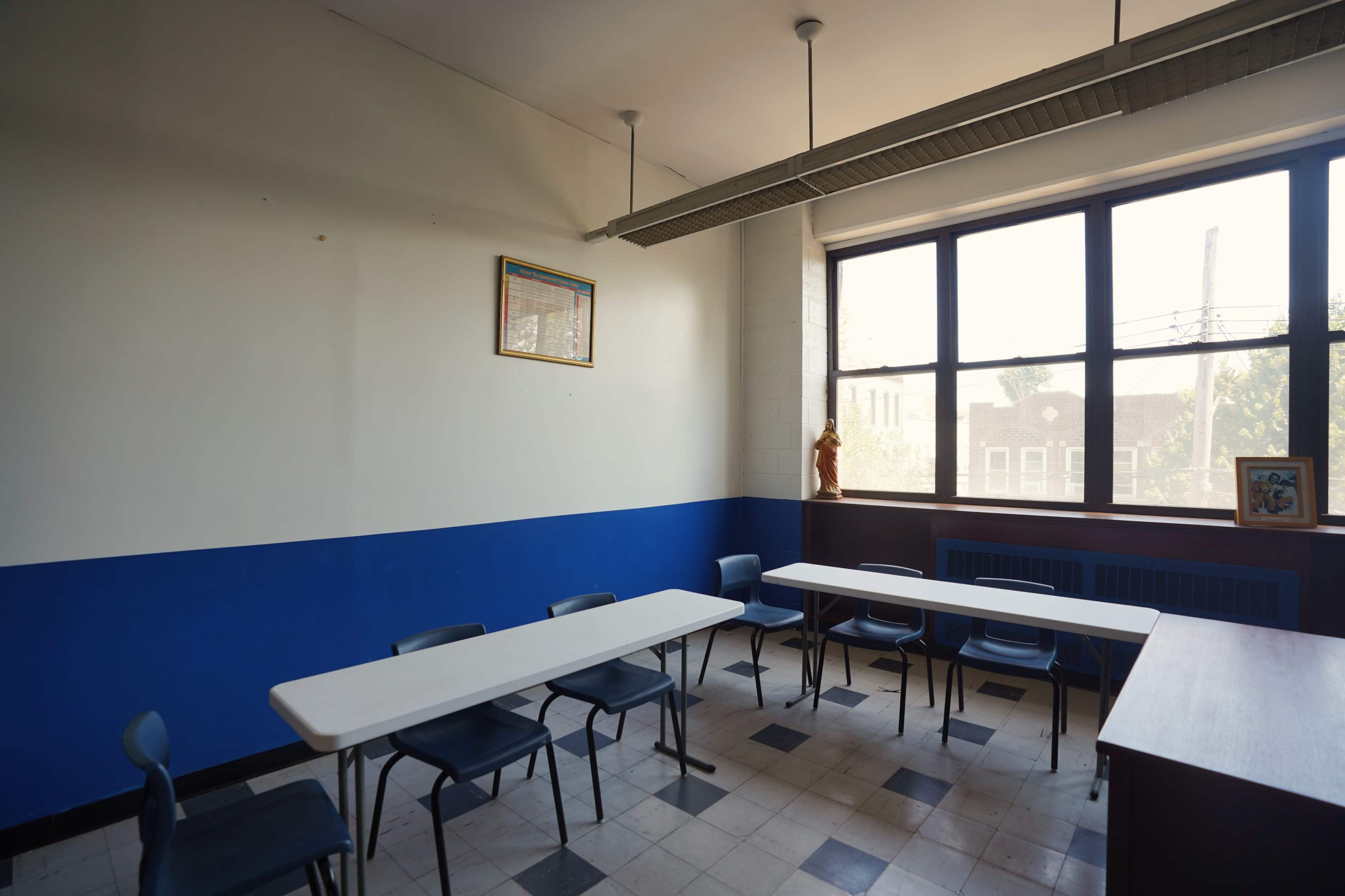 A classroom features two long tables with chairs, a blue and white color scheme, and large windows illuminating the space.