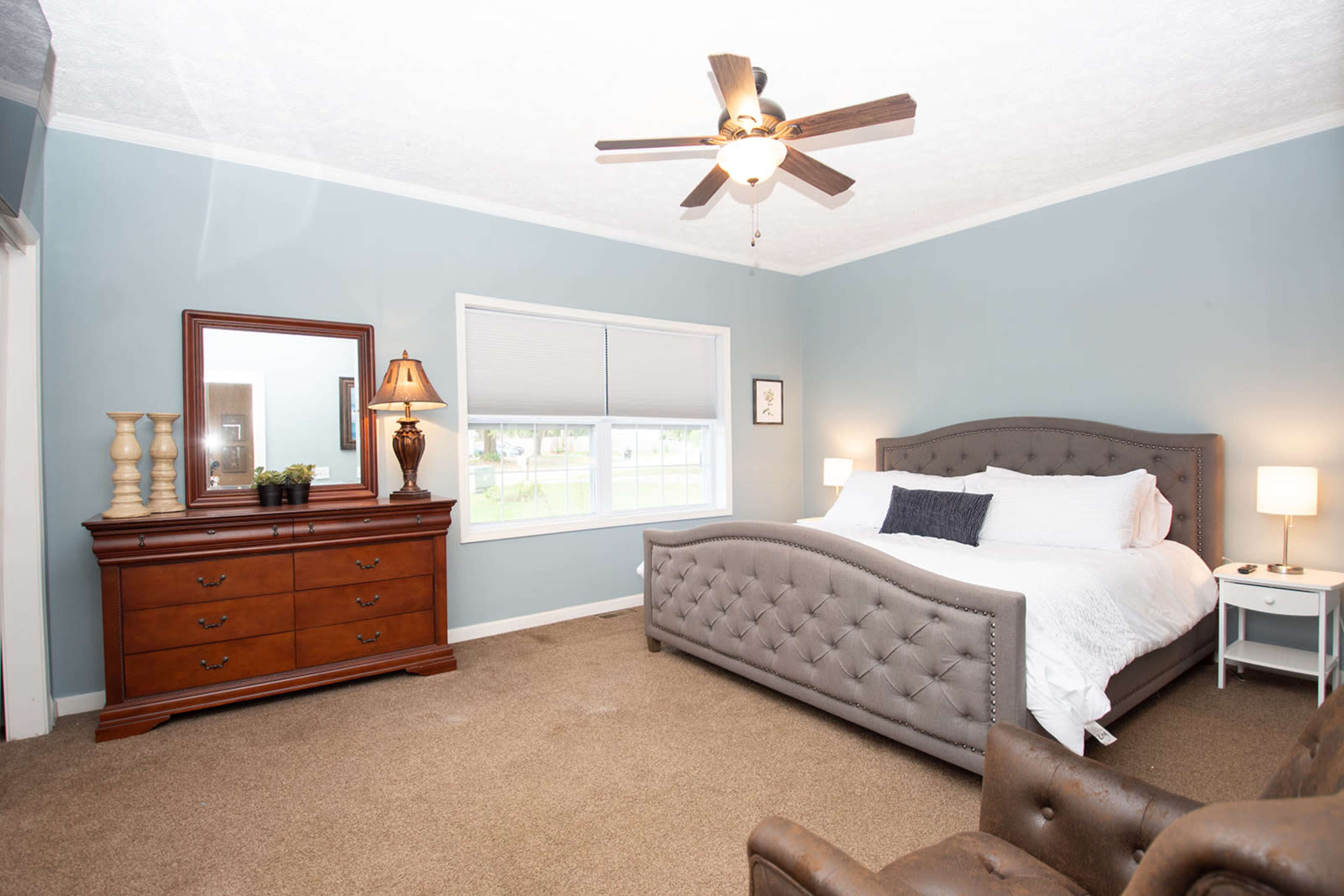 A well-furnished bedroom featuring a gray tufted bed, a wooden dresser with a lamp, and a cozy armchair.