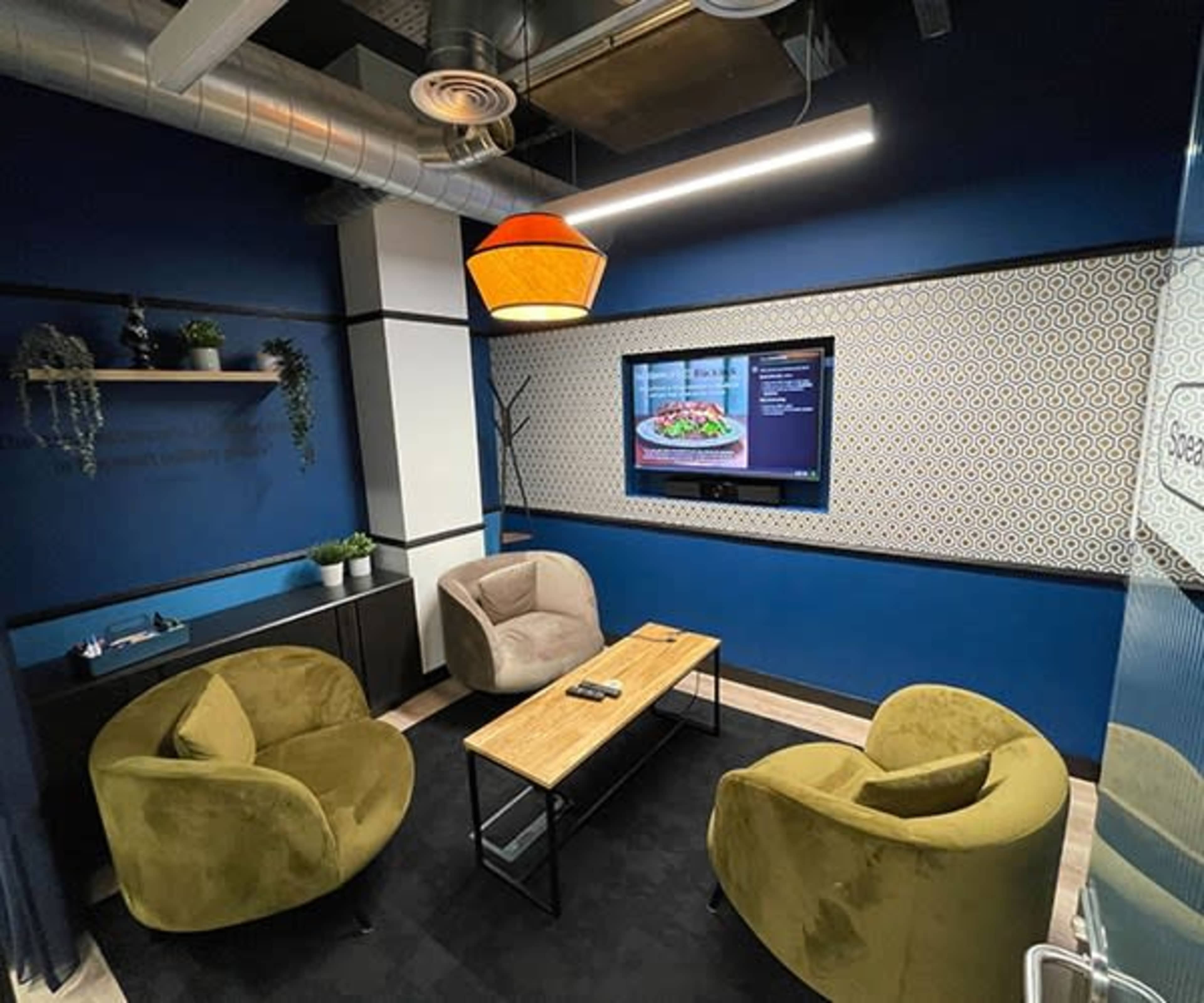 A cozy meeting room features two armchairs and a coffee table, with a television displaying a menu on a blue wall.