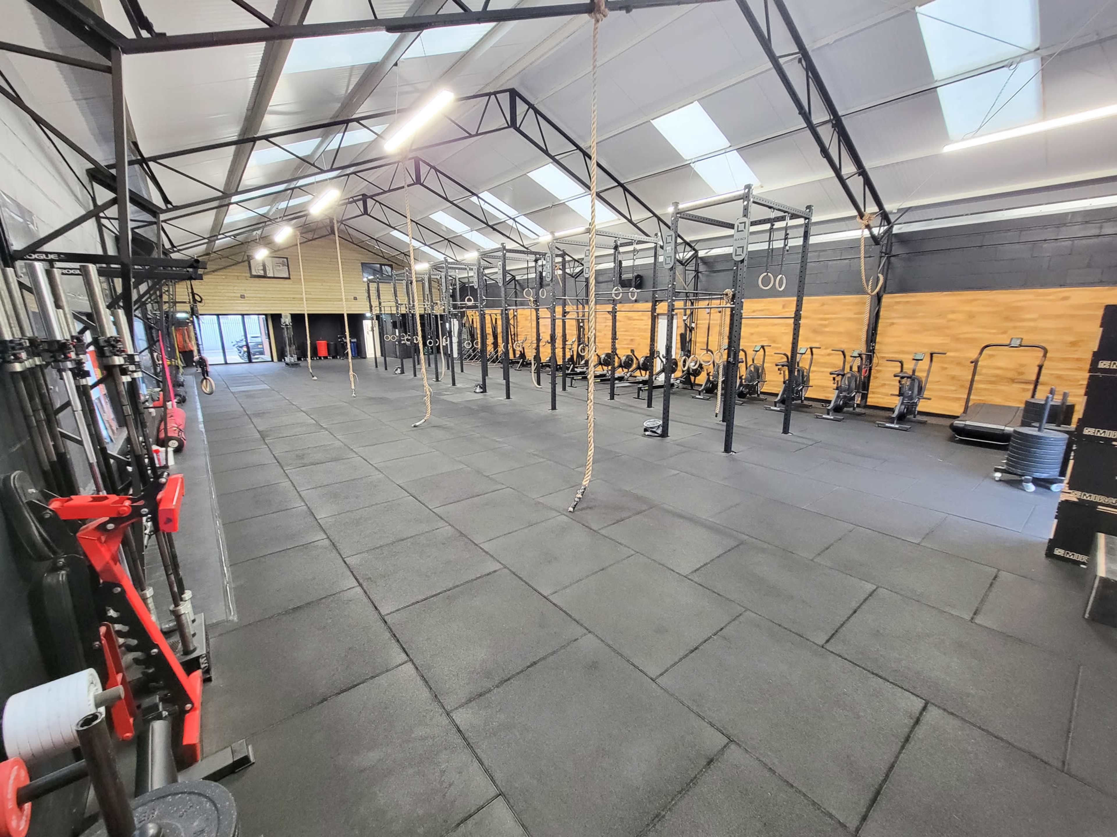 The image shows a spacious gym with exercise equipment, including pull-up bars and stationary bikes, arranged on rubber flooring under a high, vaulted ceiling.