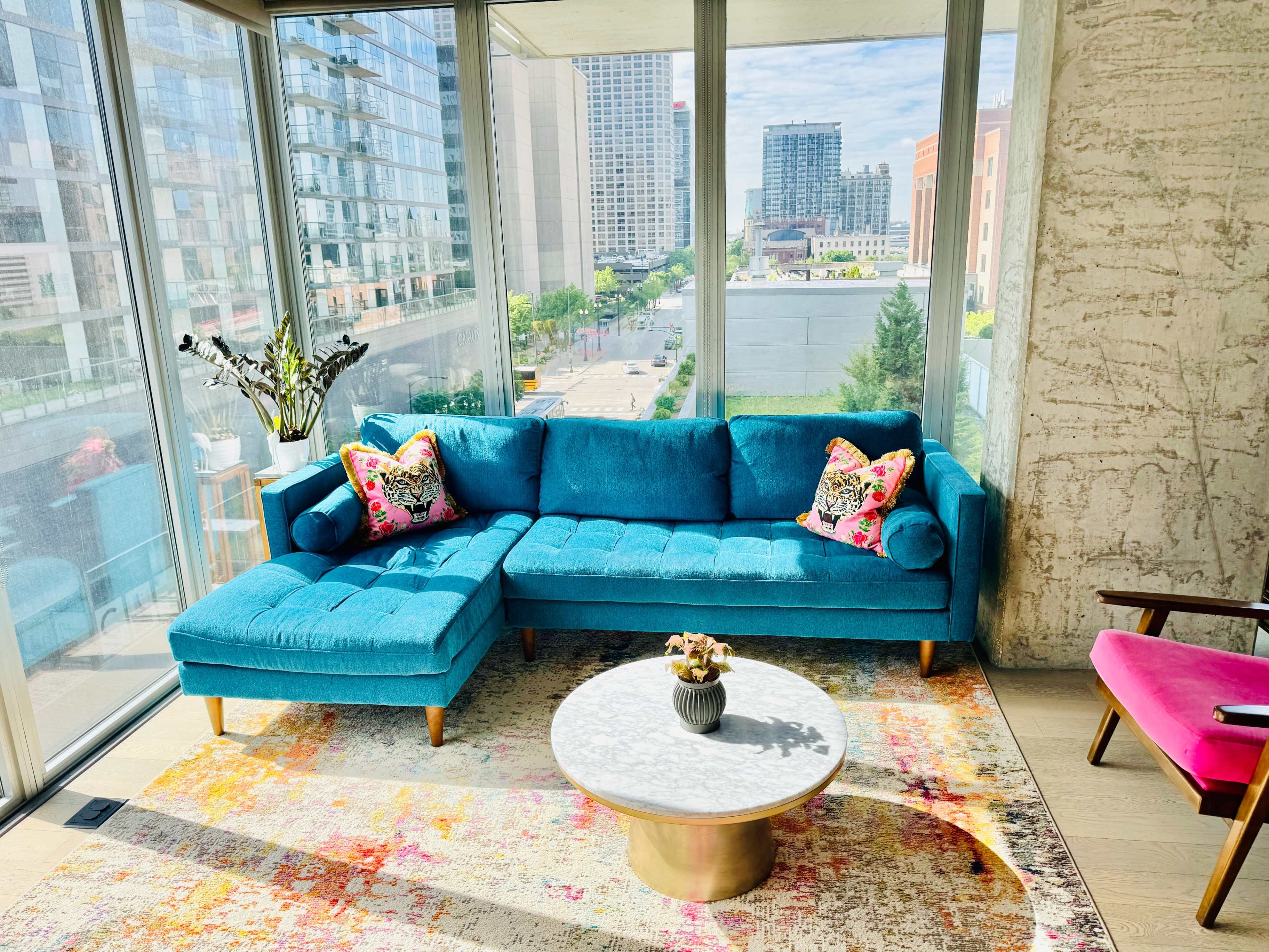 A bright living room features a turquoise sectional couch with floral accent pillows, a round coffee table, and large windows showcasing a city view.