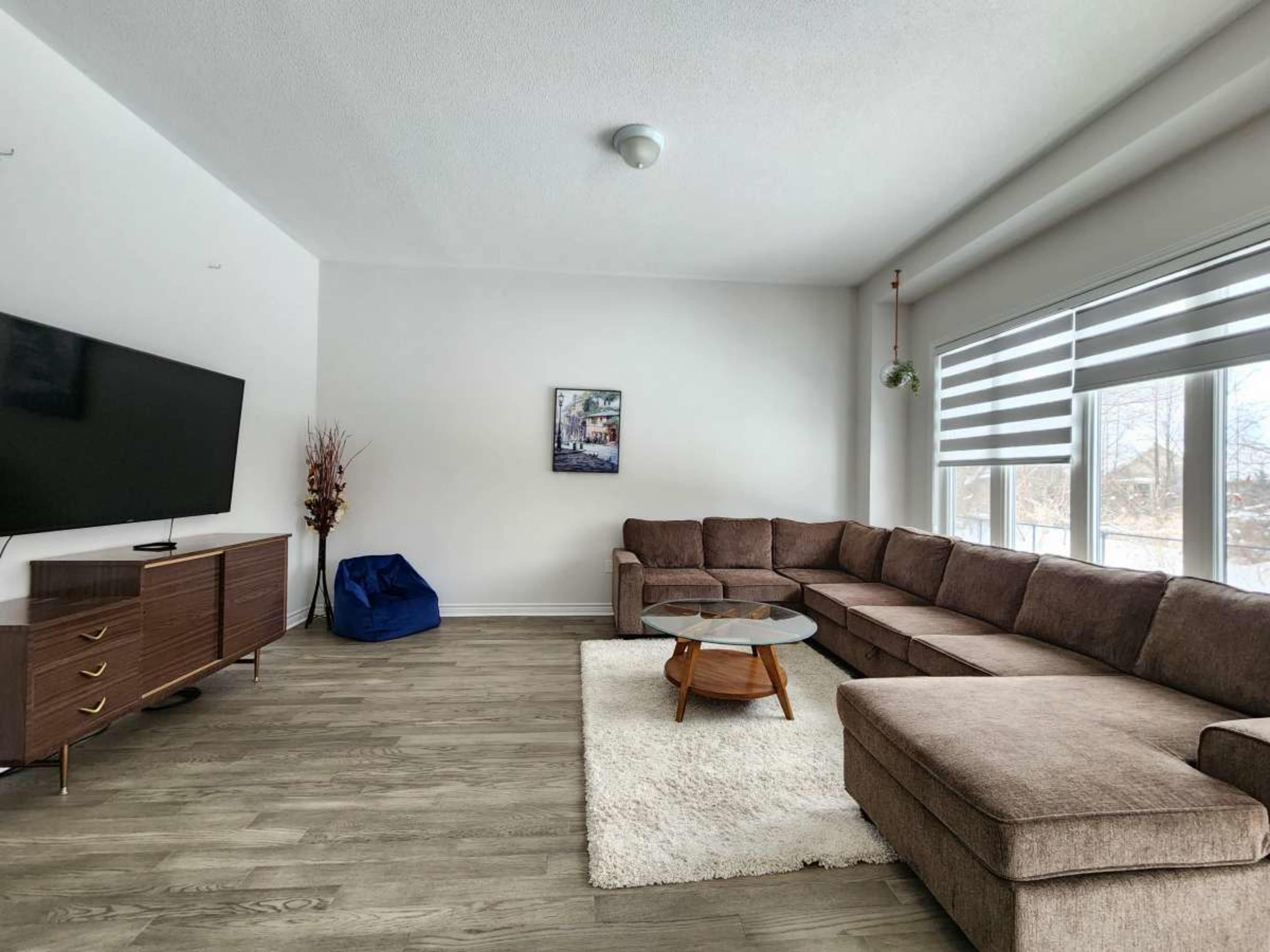 A spacious living room features a large sectional sofa, a round coffee table on a rug, a television mounted on the wall, and ample natural light from the windows.
