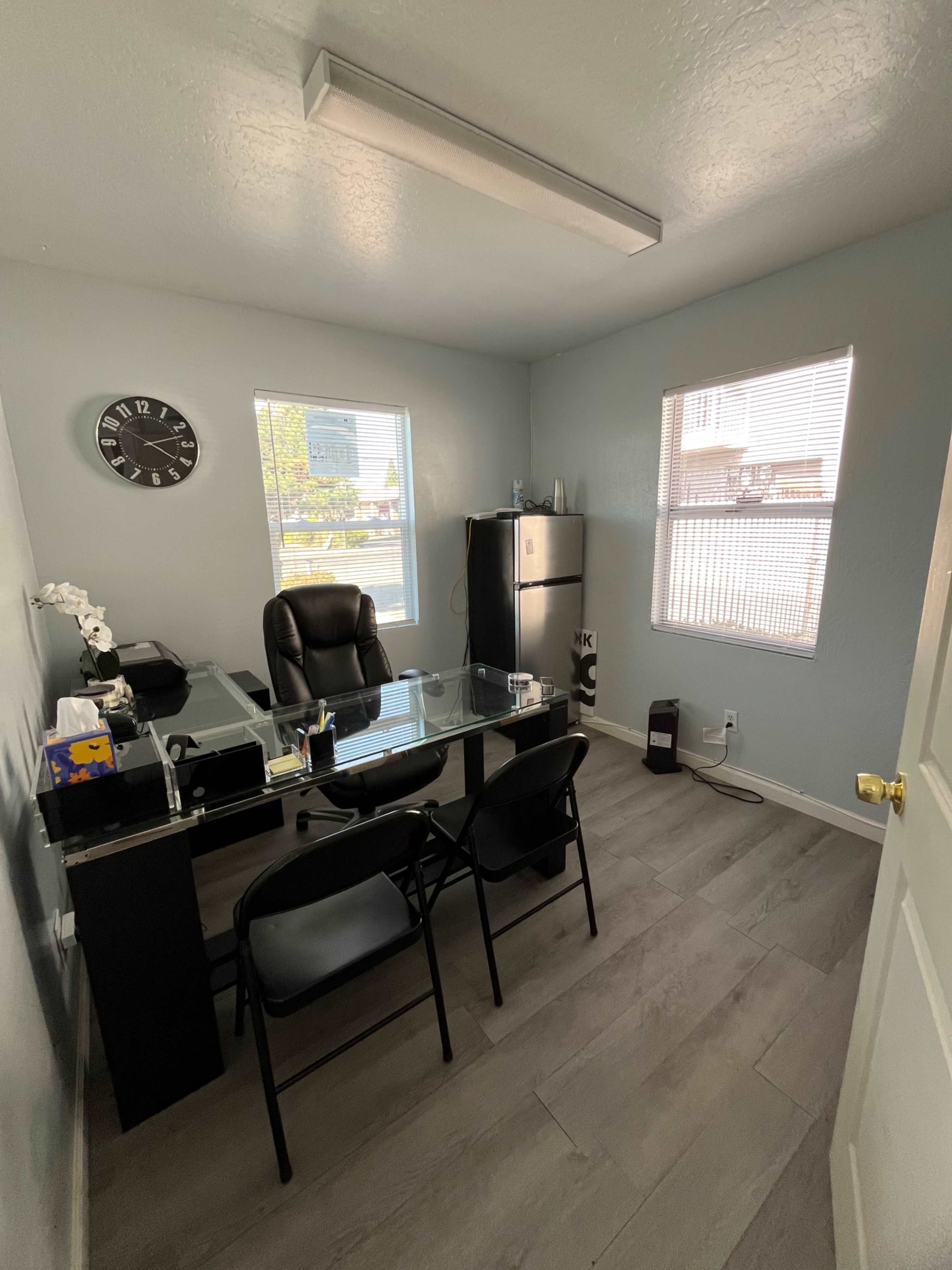 The image shows a small office with a glass desk, a black chair, two folding chairs, a refrigerator, and large windows allowing natural light.