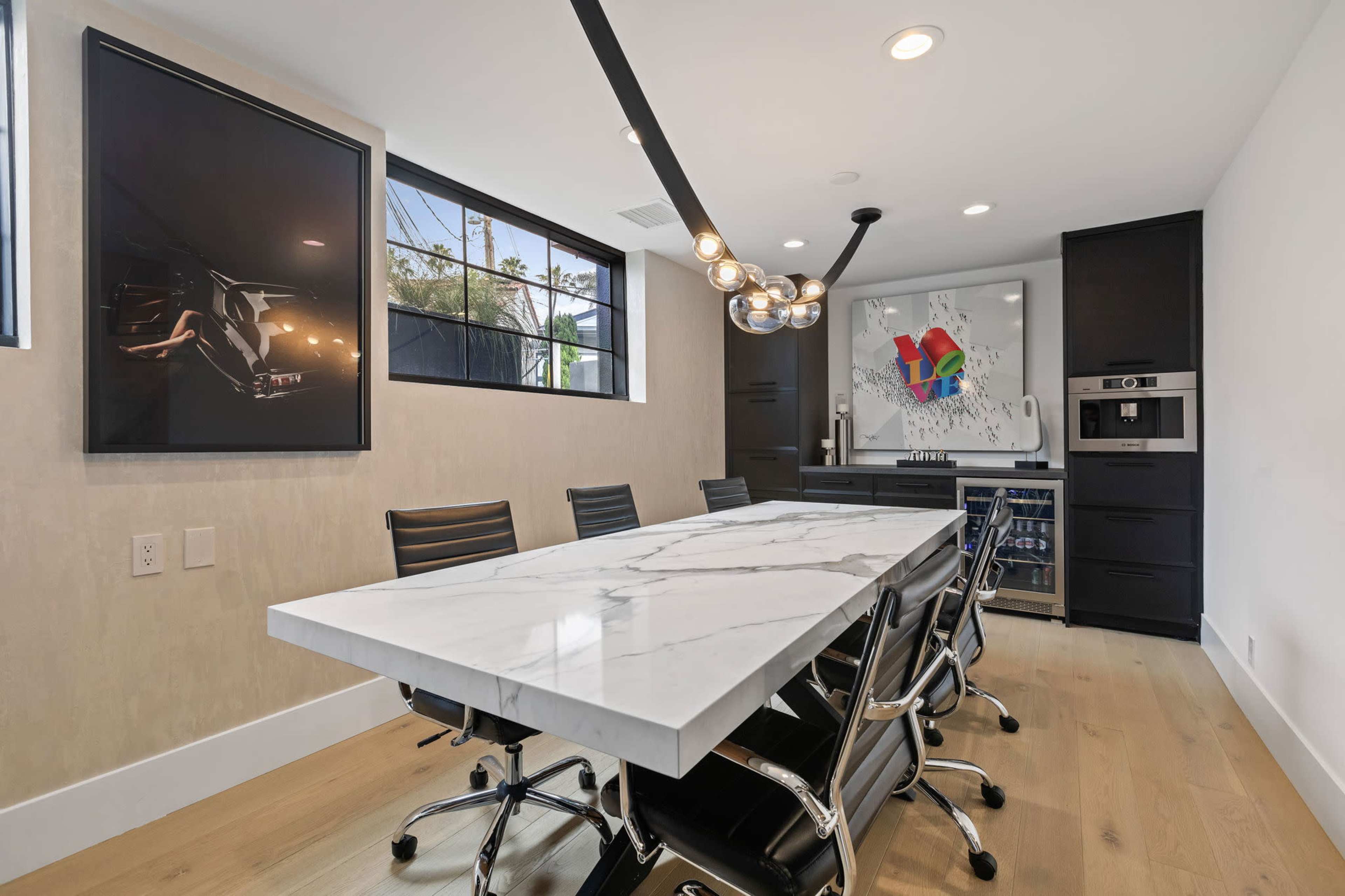 A modern dining area features a long marble table surrounded by black chairs, with large windows and artwork on the walls.