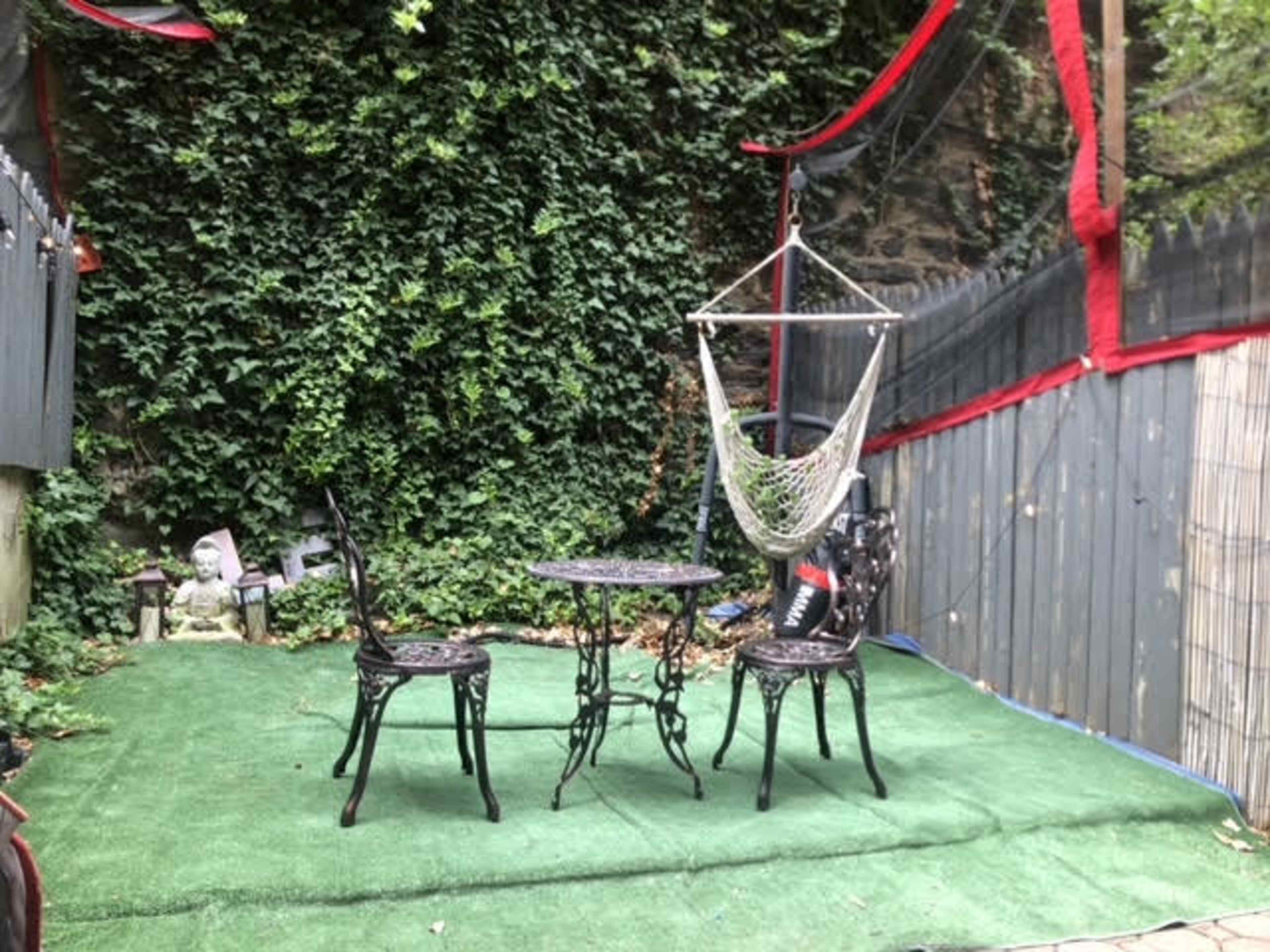 A small outdoor space with two chairs, a round table, a hammock, and a green carpeted area surrounded by ivy-covered walls.