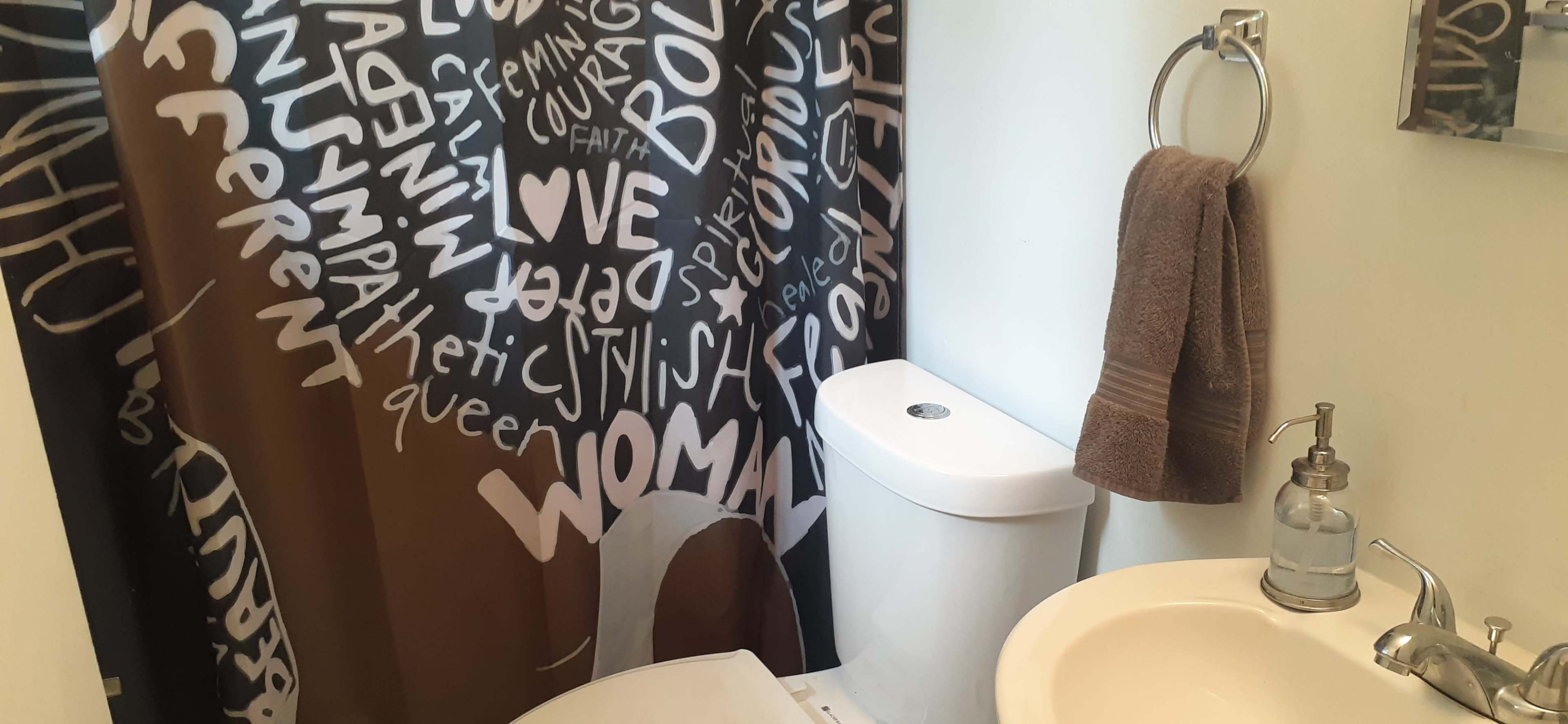 A small bathroom with a dark shower curtain featuring white text, a toilet, a sink with a soap dispenser, and a towel hanging on a rack.