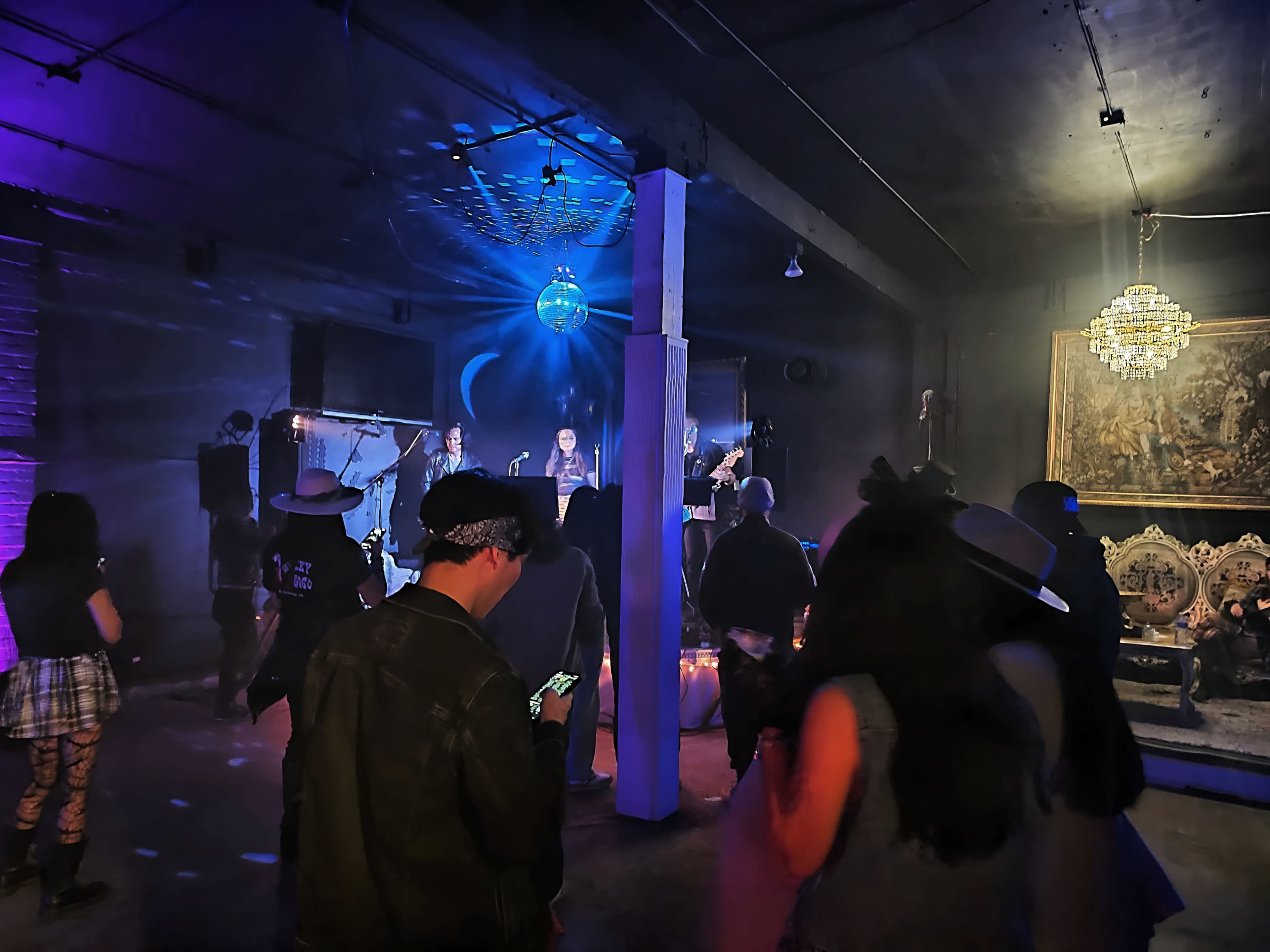A band performs on stage while people dance and socialize in a dimly lit venue adorned with decorative lighting.