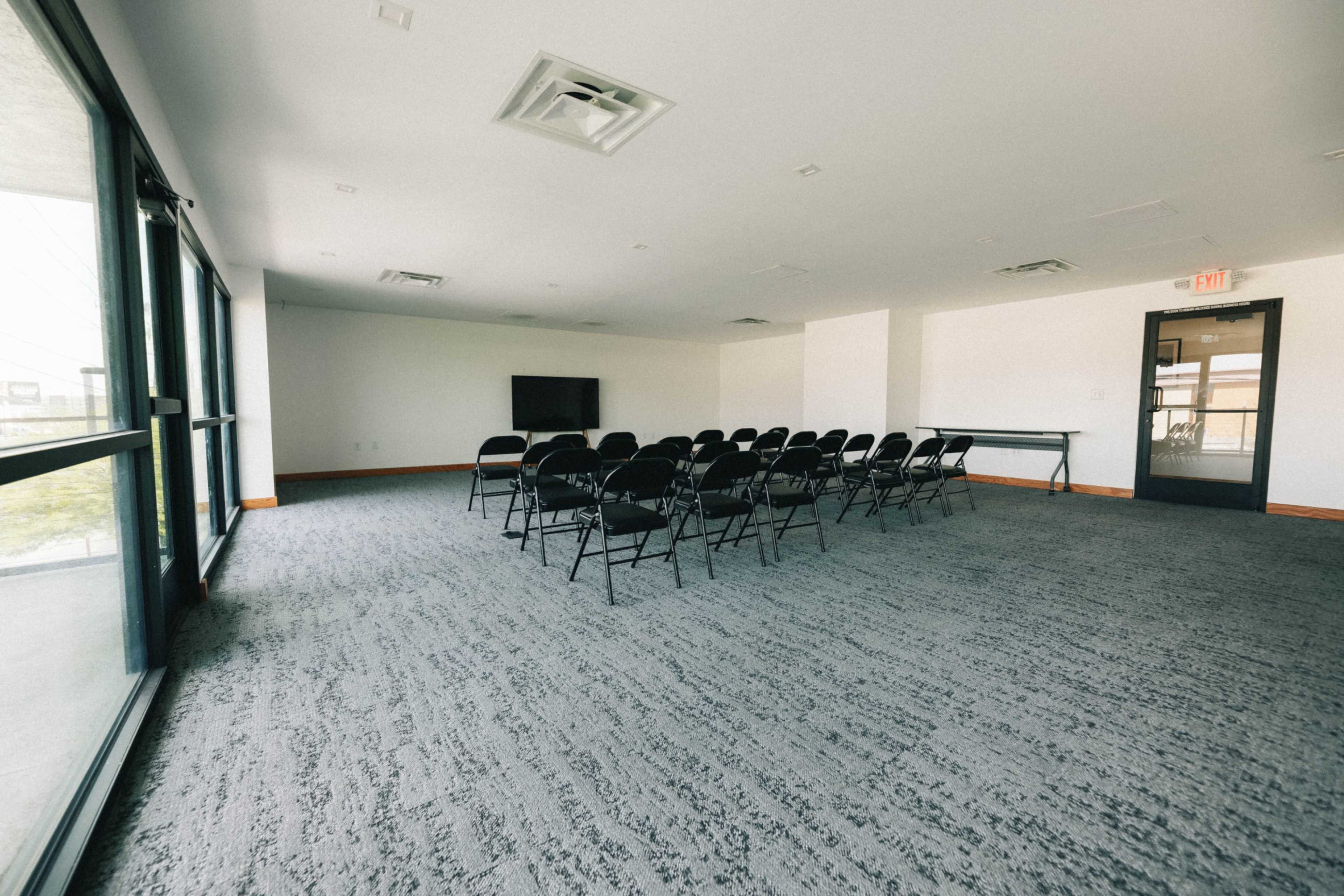 The image shows an empty conference room with rows of black chairs facing a wall-mounted television and large windows.