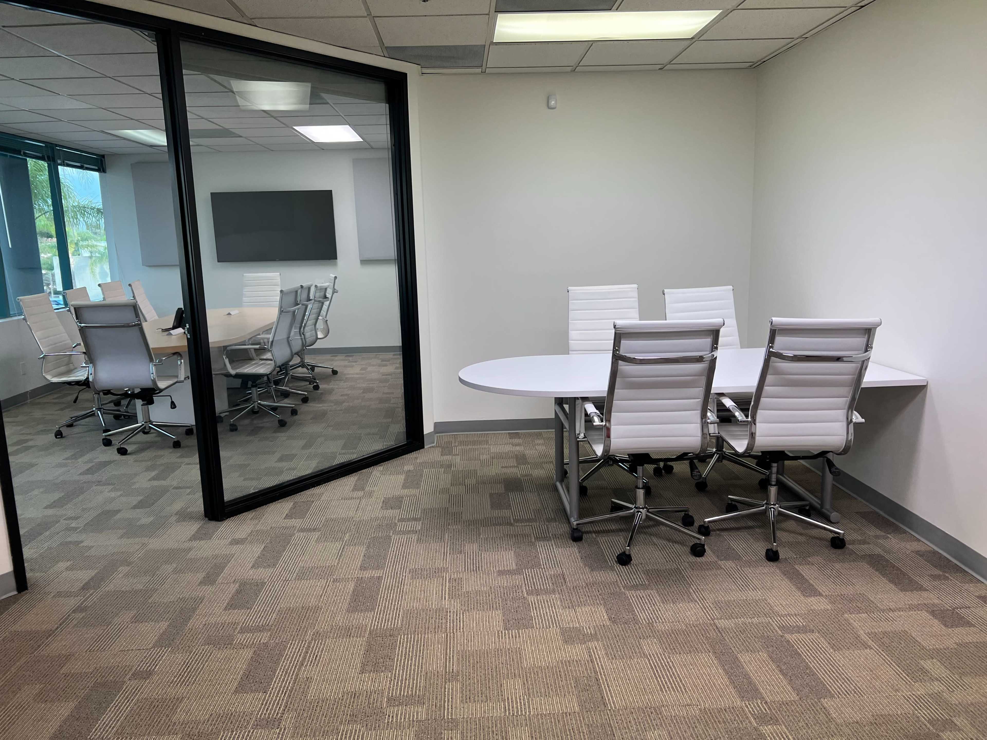 A modern office space with a glass-walled conference room and a separate table with white chairs in an adjacent area.