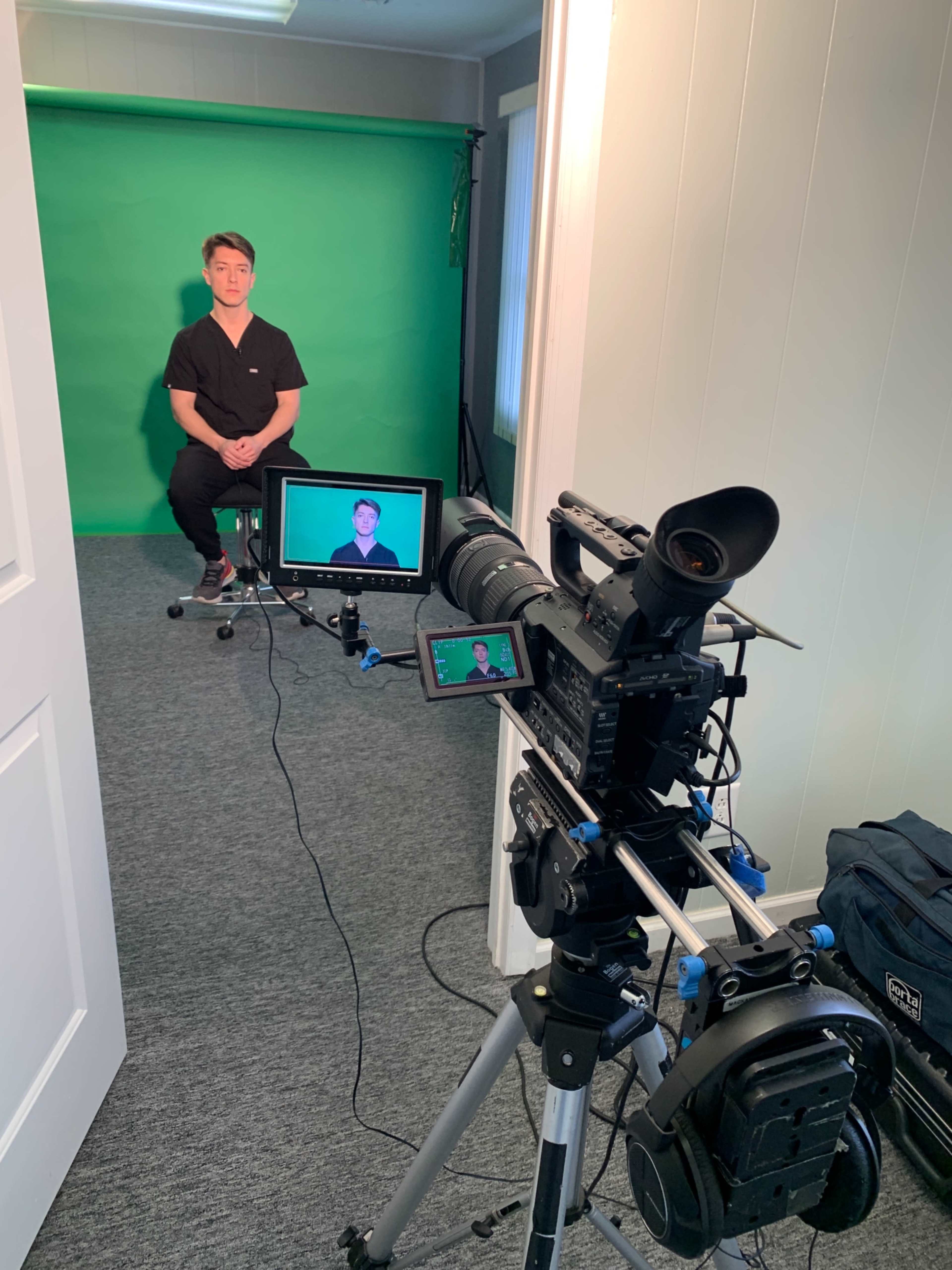 A person sits on a stool in front of a green screen while being filmed by a camera on a tripod.