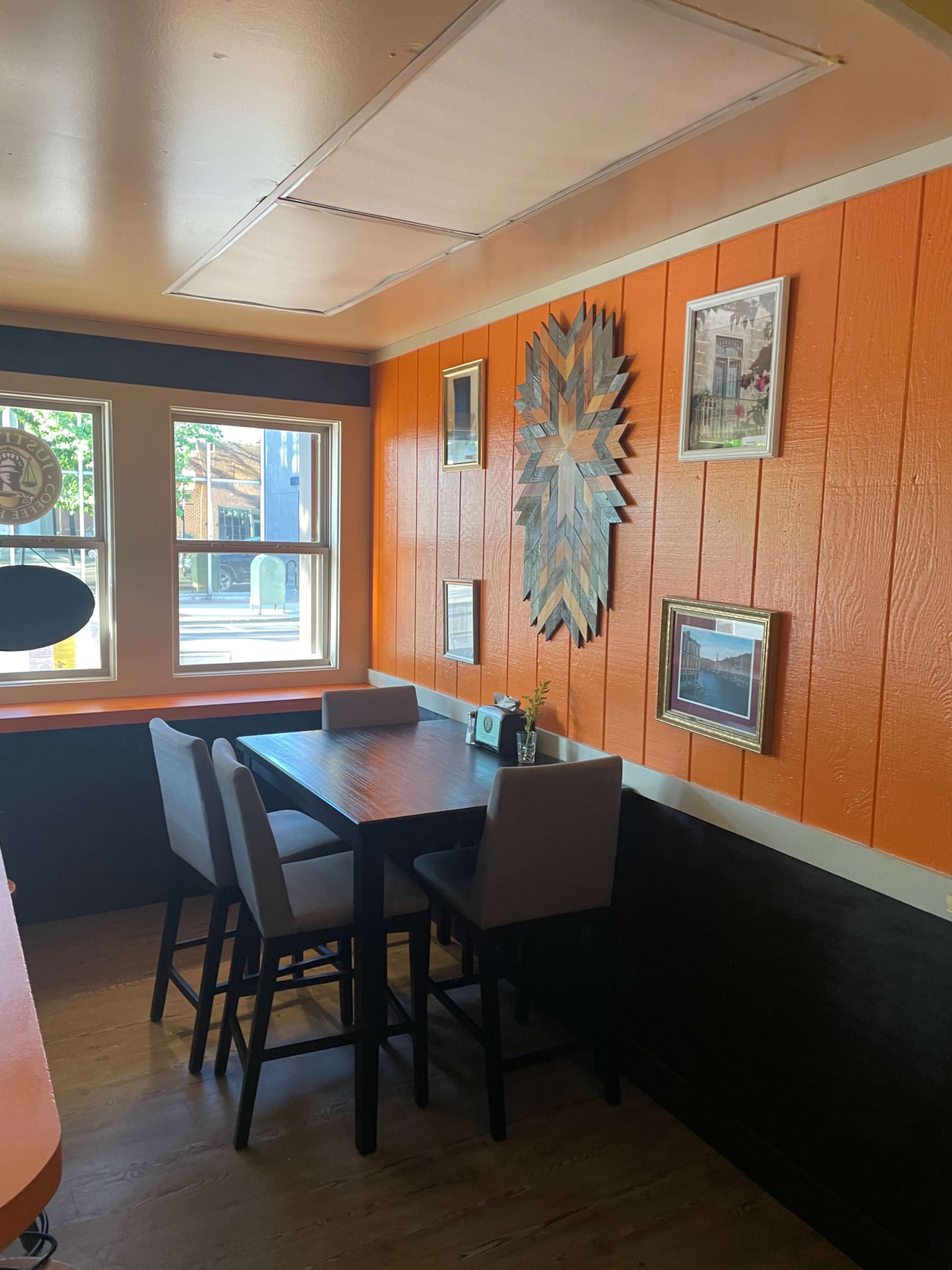A cozy dining nook with a wooden table and four chairs against an orange and dark wood paneled wall, adorned with various framed pictures and decorations.