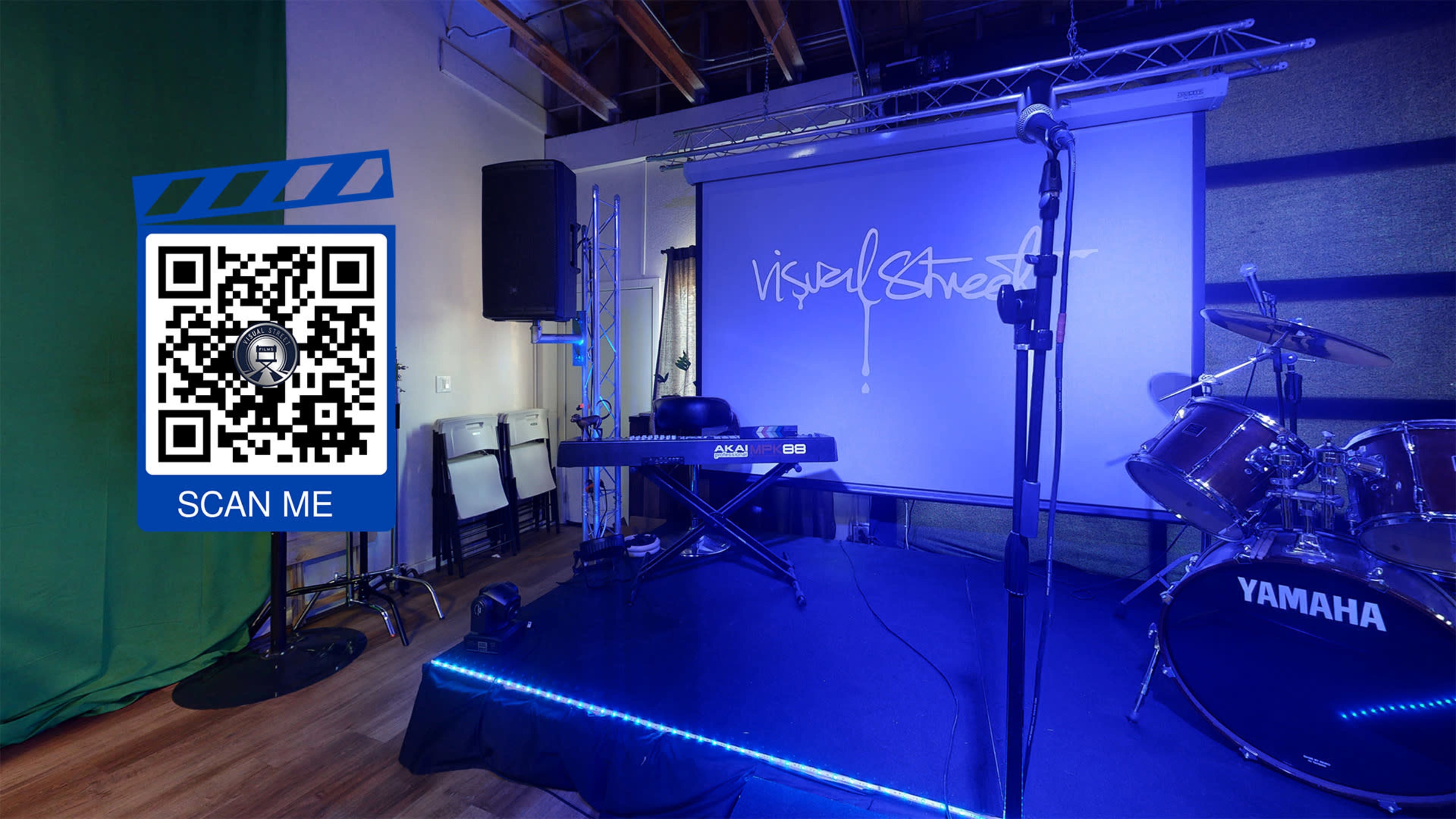 A small stage setup featuring a keyboard, a drum set, and a projector screen displaying the text "Visually Street," along with a QR code and a green backdrop.