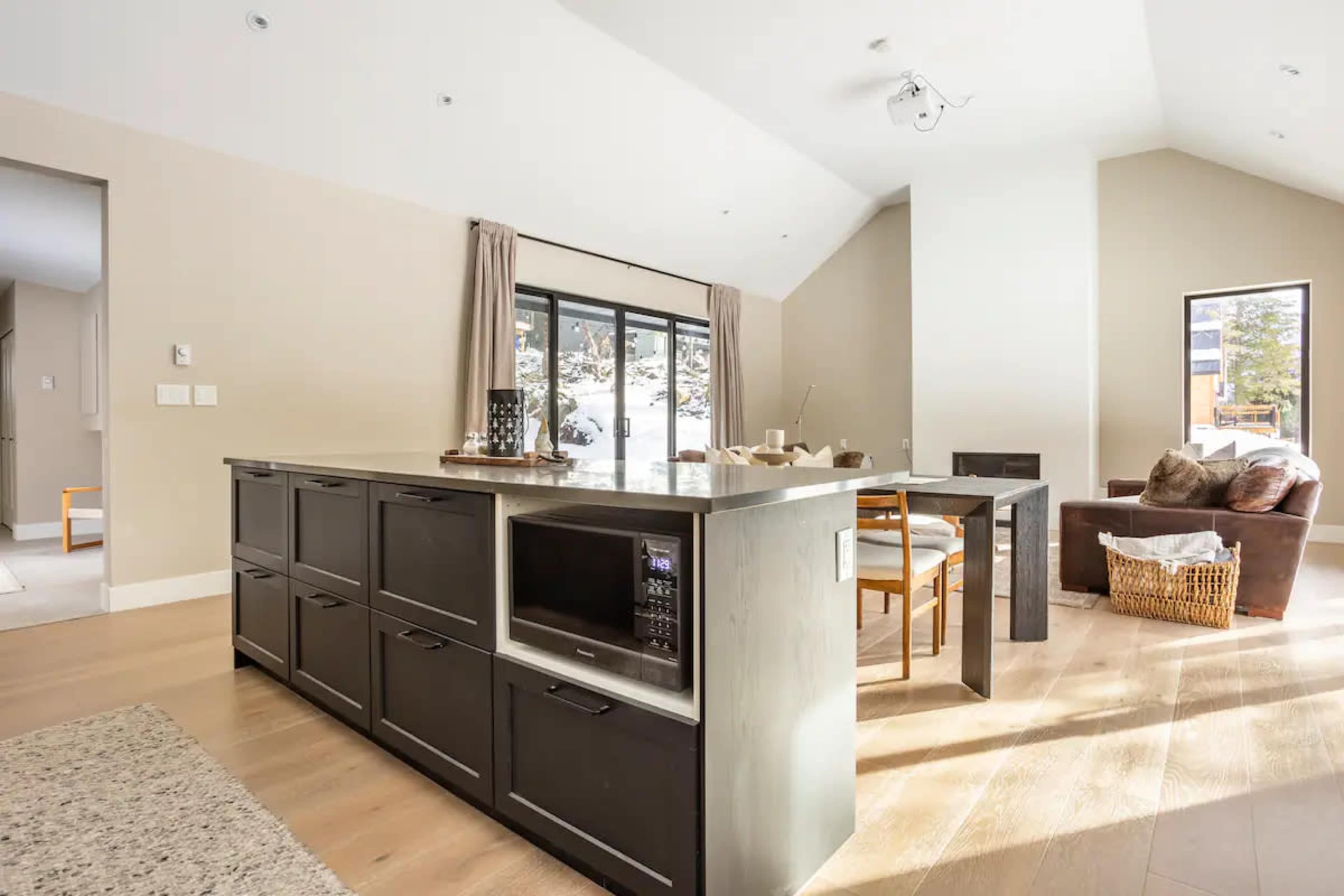 A modern kitchen with an island featuring a built-in microwave, surrounded by a dining area with a table and chairs, and a living space with large windows.