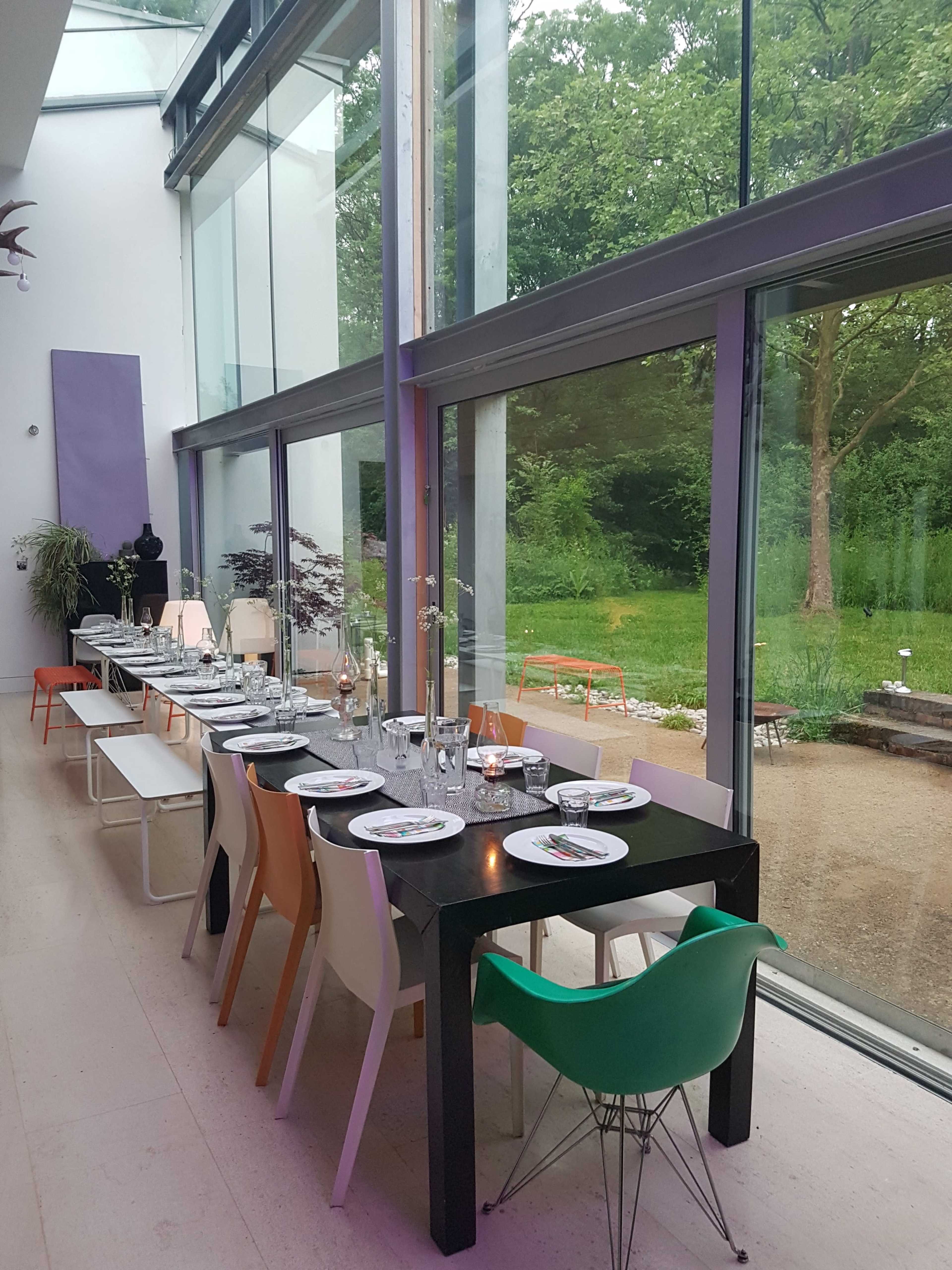 A long dining table is set with plates and glassware in a modern room featuring large windows that overlook a green garden.