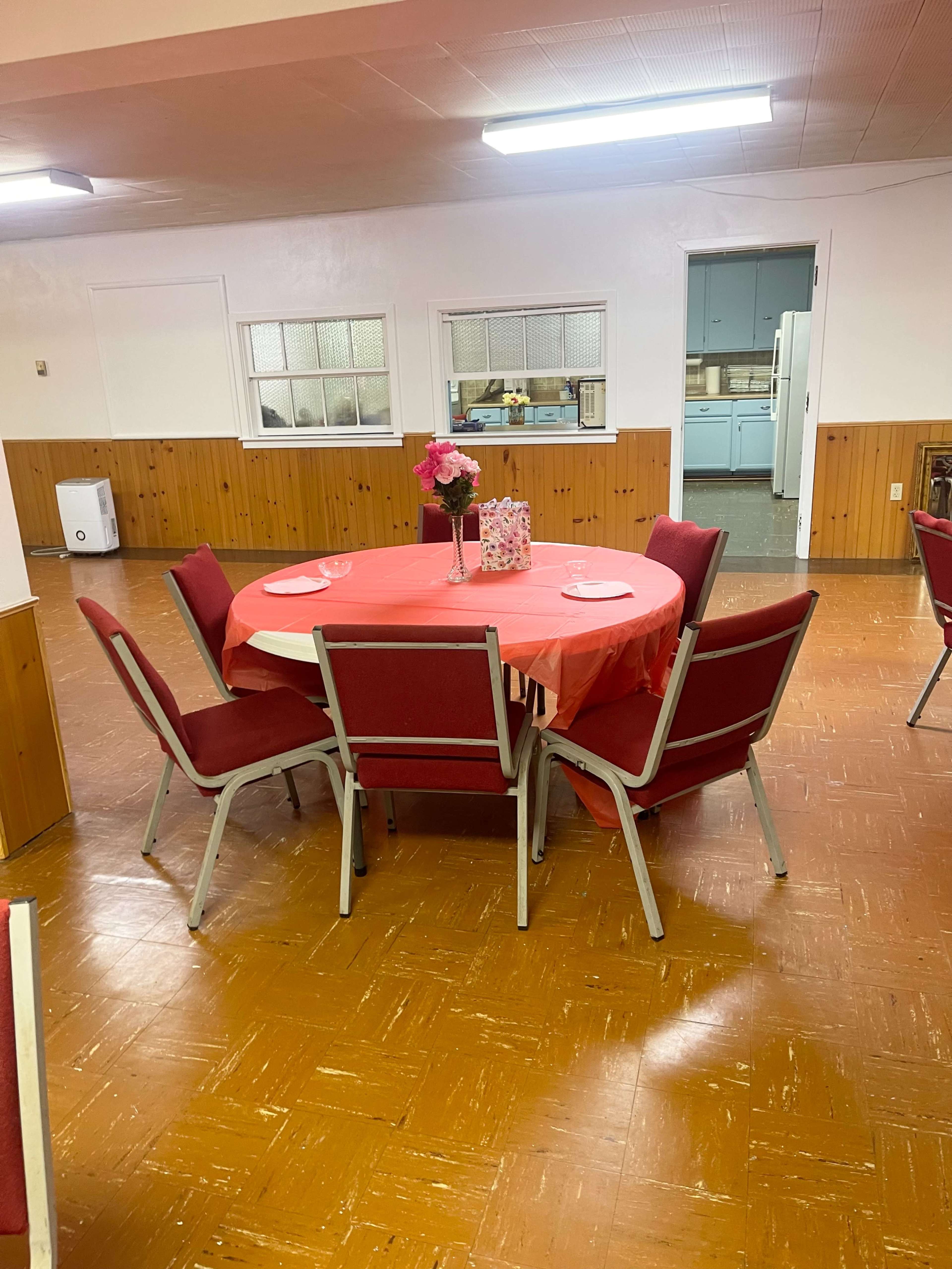 A round table covered with a red cloth is set in a sparsely decorated room with wooden paneling and several red chairs arranged around it.