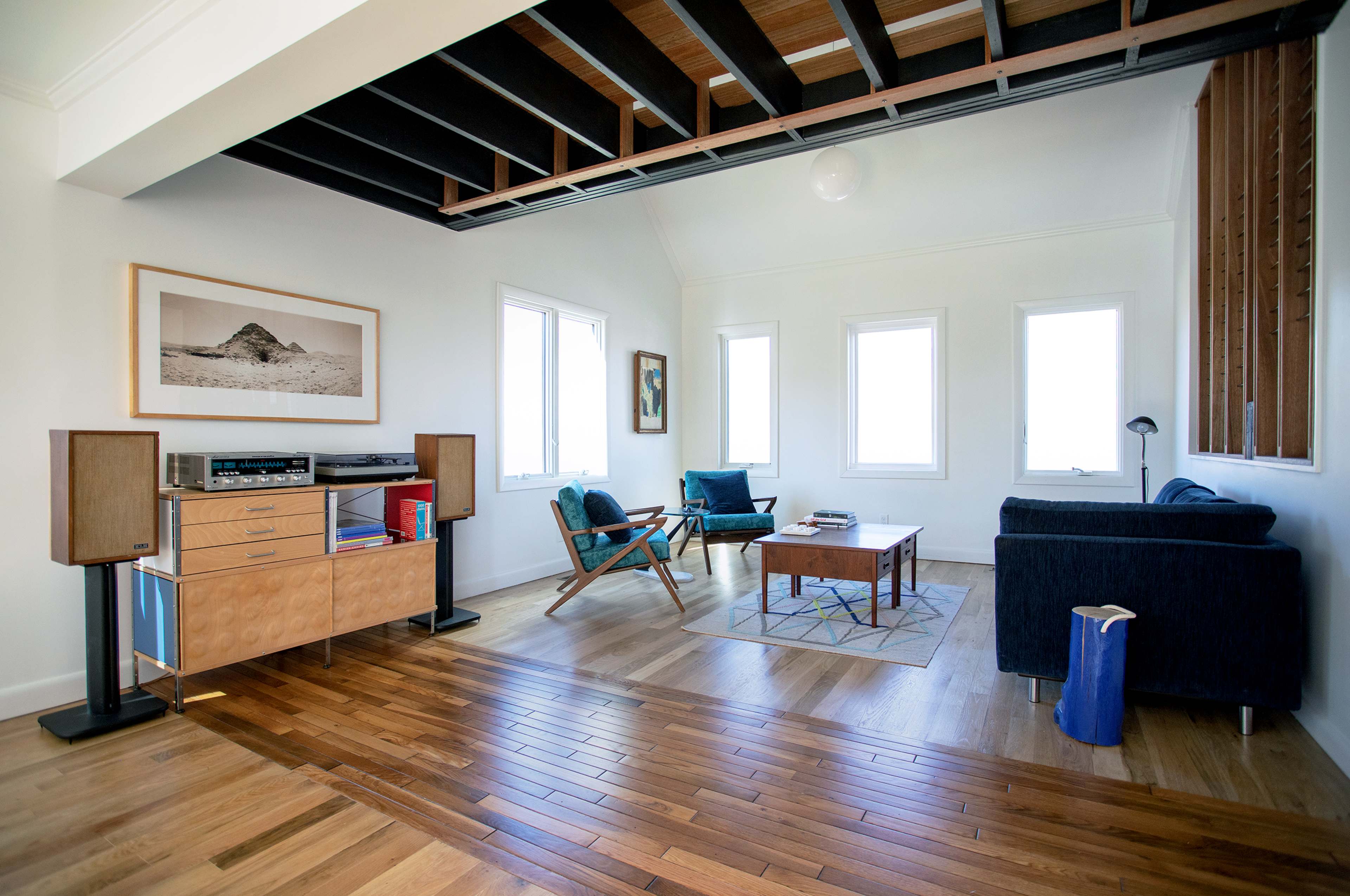 A modern living room with wooden floors, a coffee table, blue chairs, and large windows.