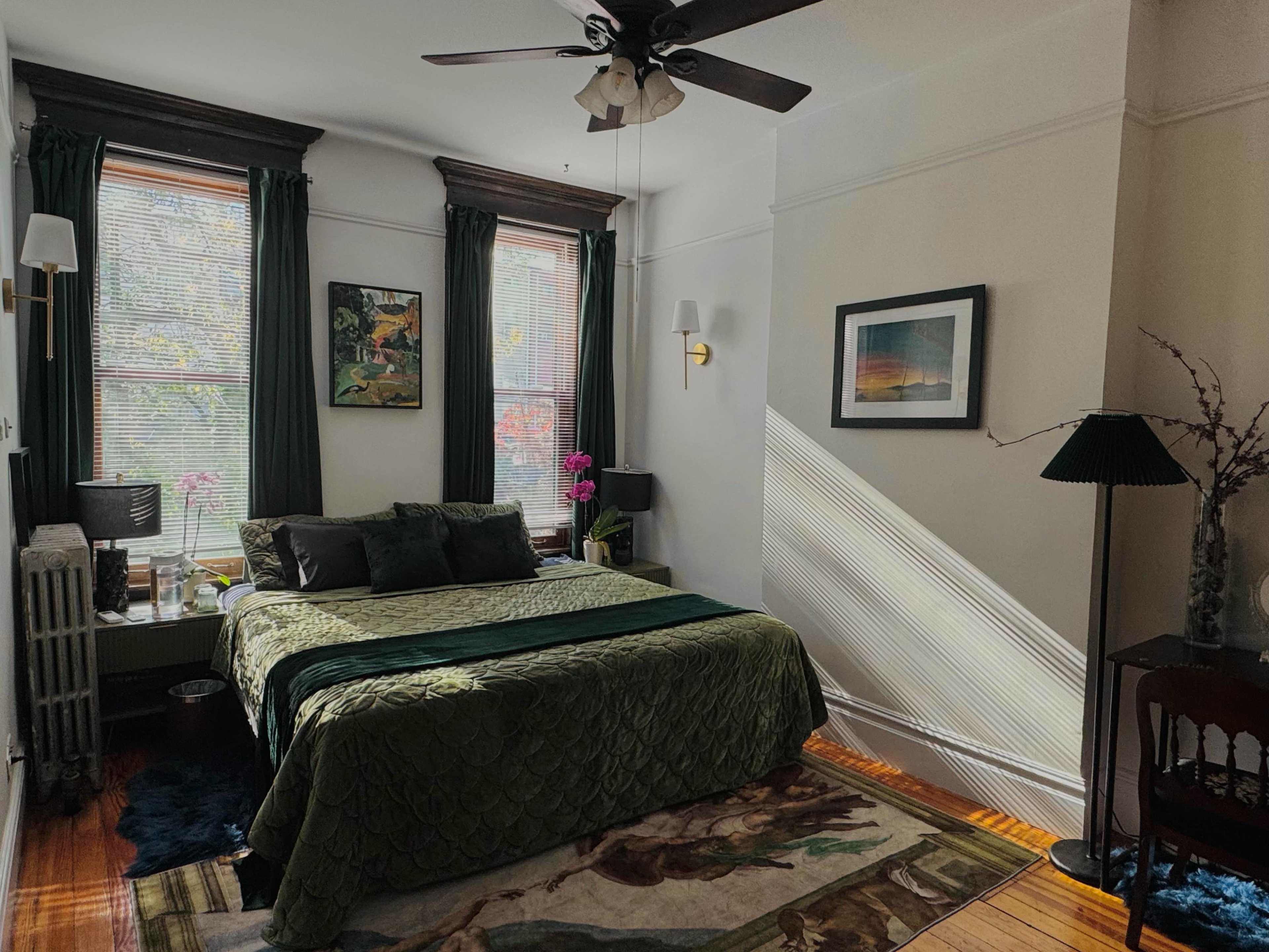 The image shows a cozy bedroom featuring a green quilted bedspread, two windows with dark curtains, and a mix of decorative elements including a painting and a table lamp.