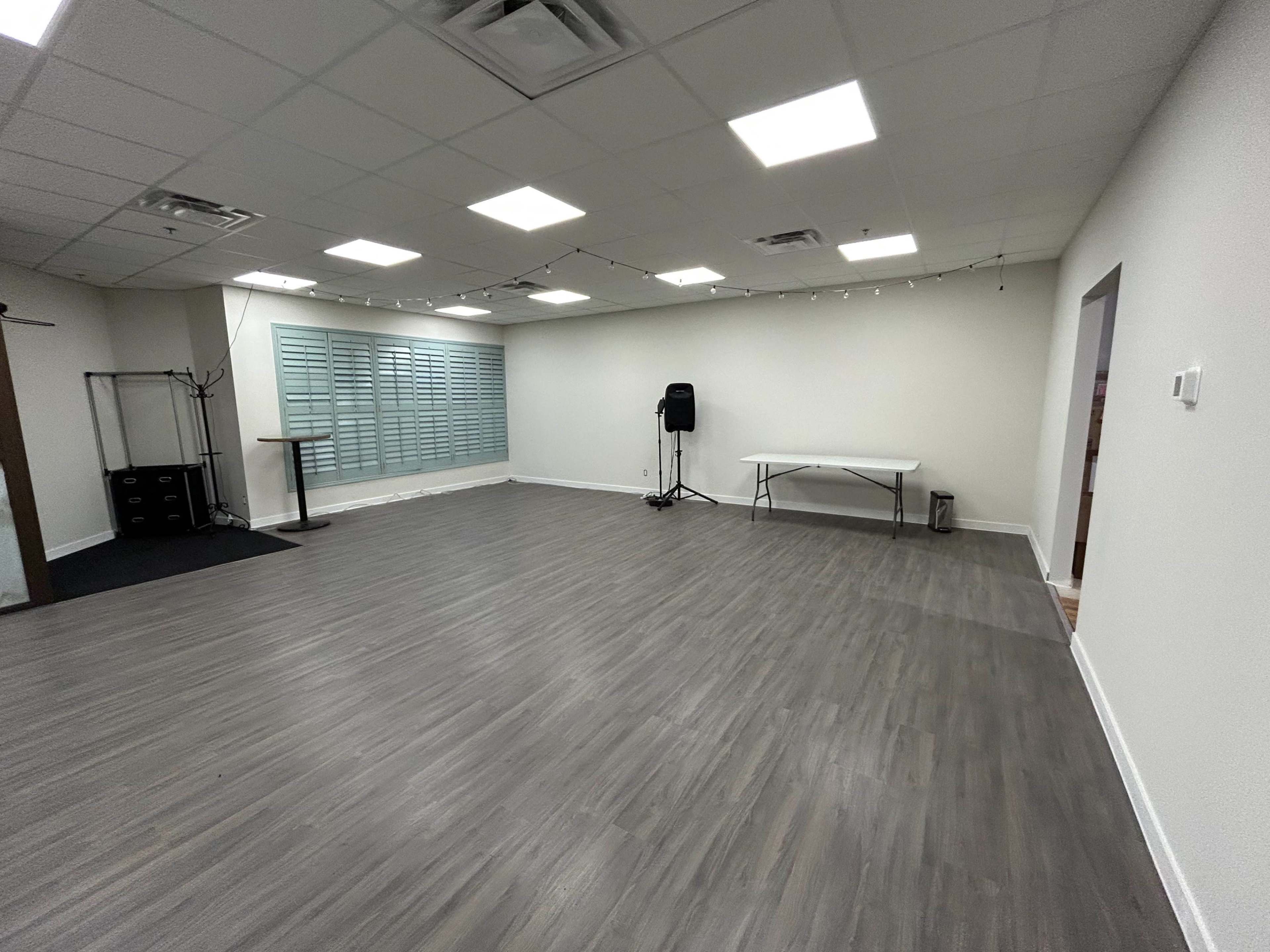 The image shows a spacious, empty room with a gray wooden floor, overhead lighting, and a few pieces of furniture including a table and a speaker.